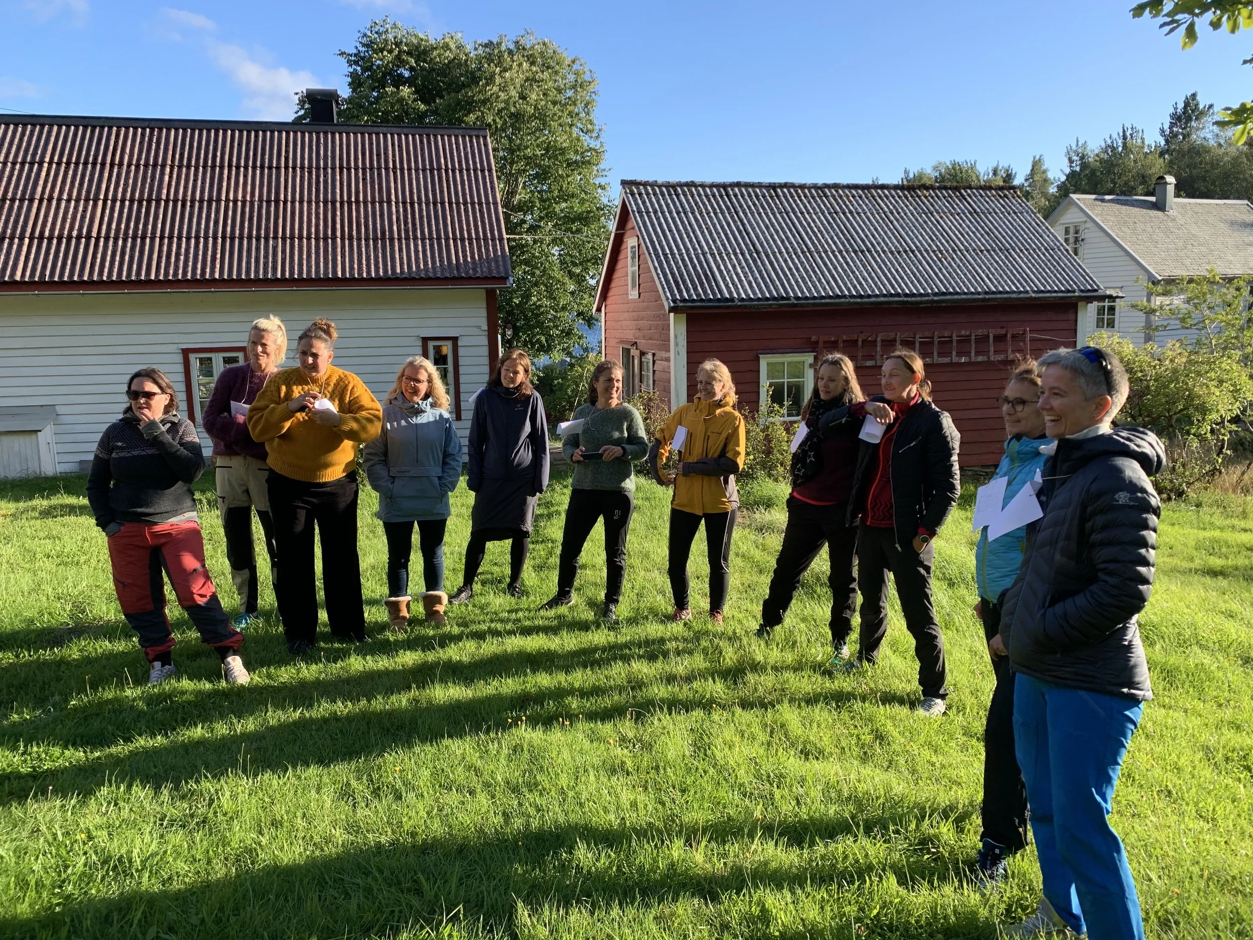 En gruppe mennesker står samlet utendørs på gresset med hus i bakgrunnen på en solrik dag.