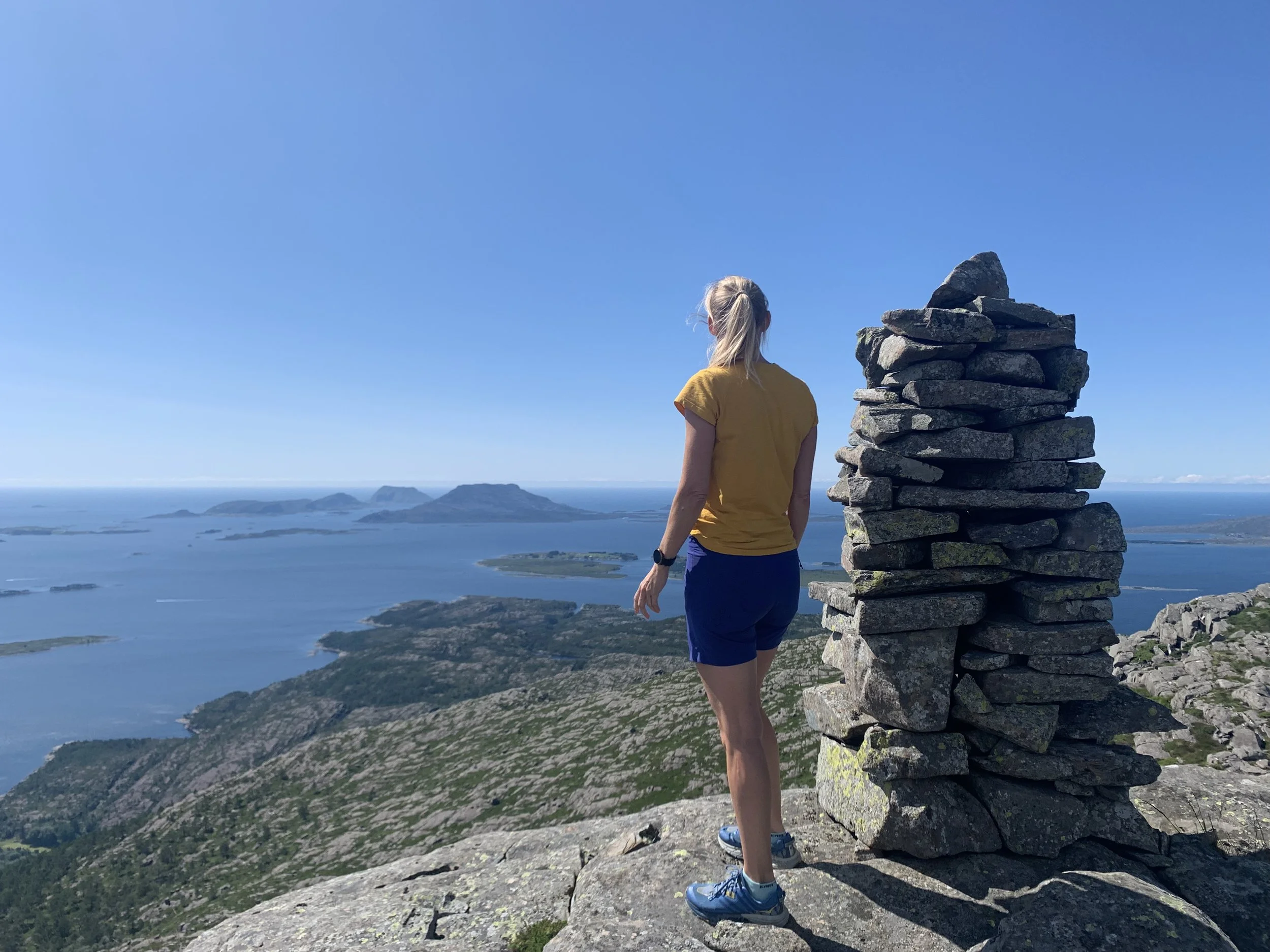 En person står på fjelltopp og ser utover sjø og øyer, med en lav steinpyramide ved siden av. Det er klart vær med blå himmel.