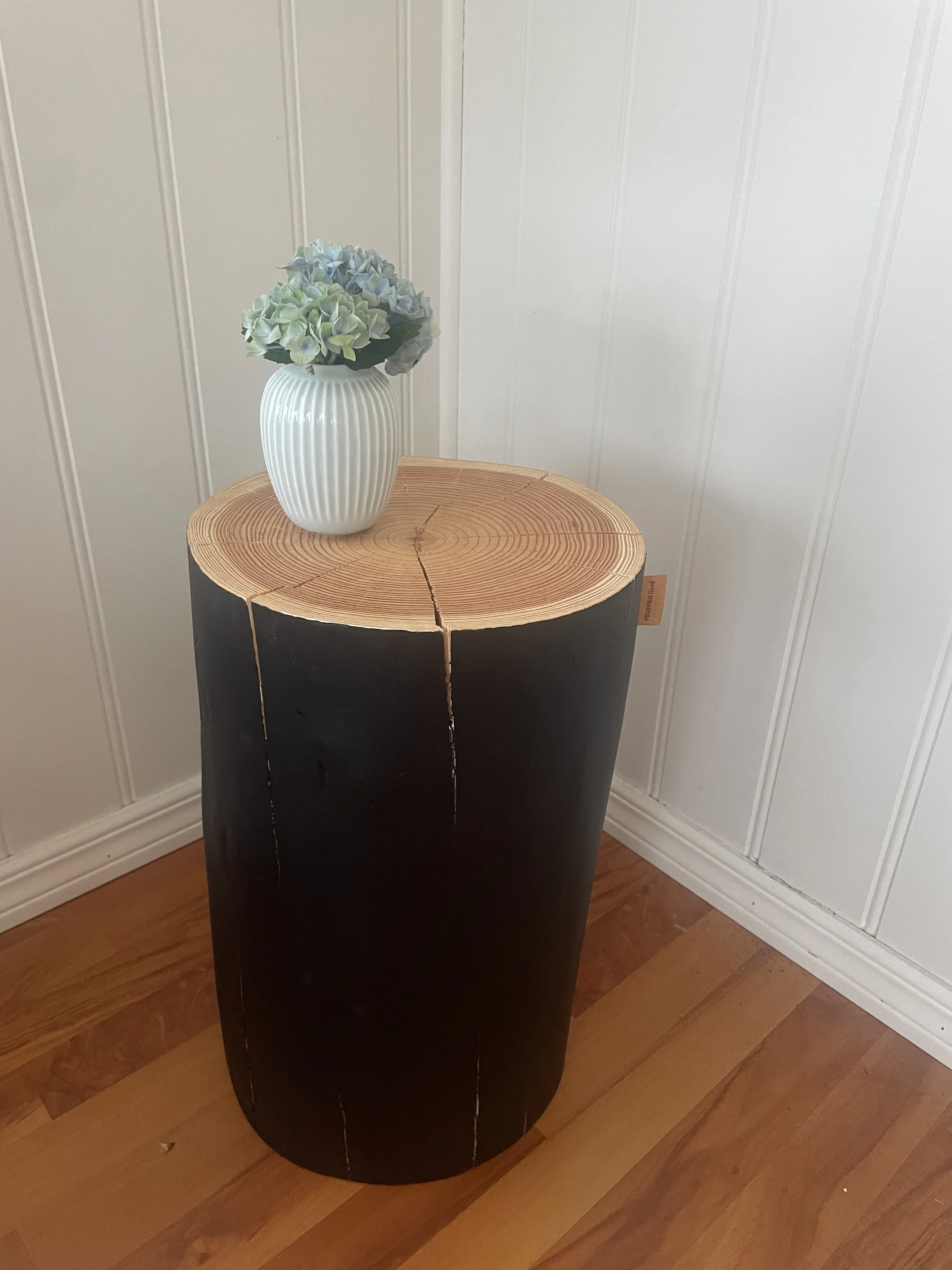 Et rund svart trebord med en tynn kubbe av tre i midten, på toppen står en hvit vase med falmet blå og lilla hortensia blomster foran en hvit vegg med vertikale paneler.