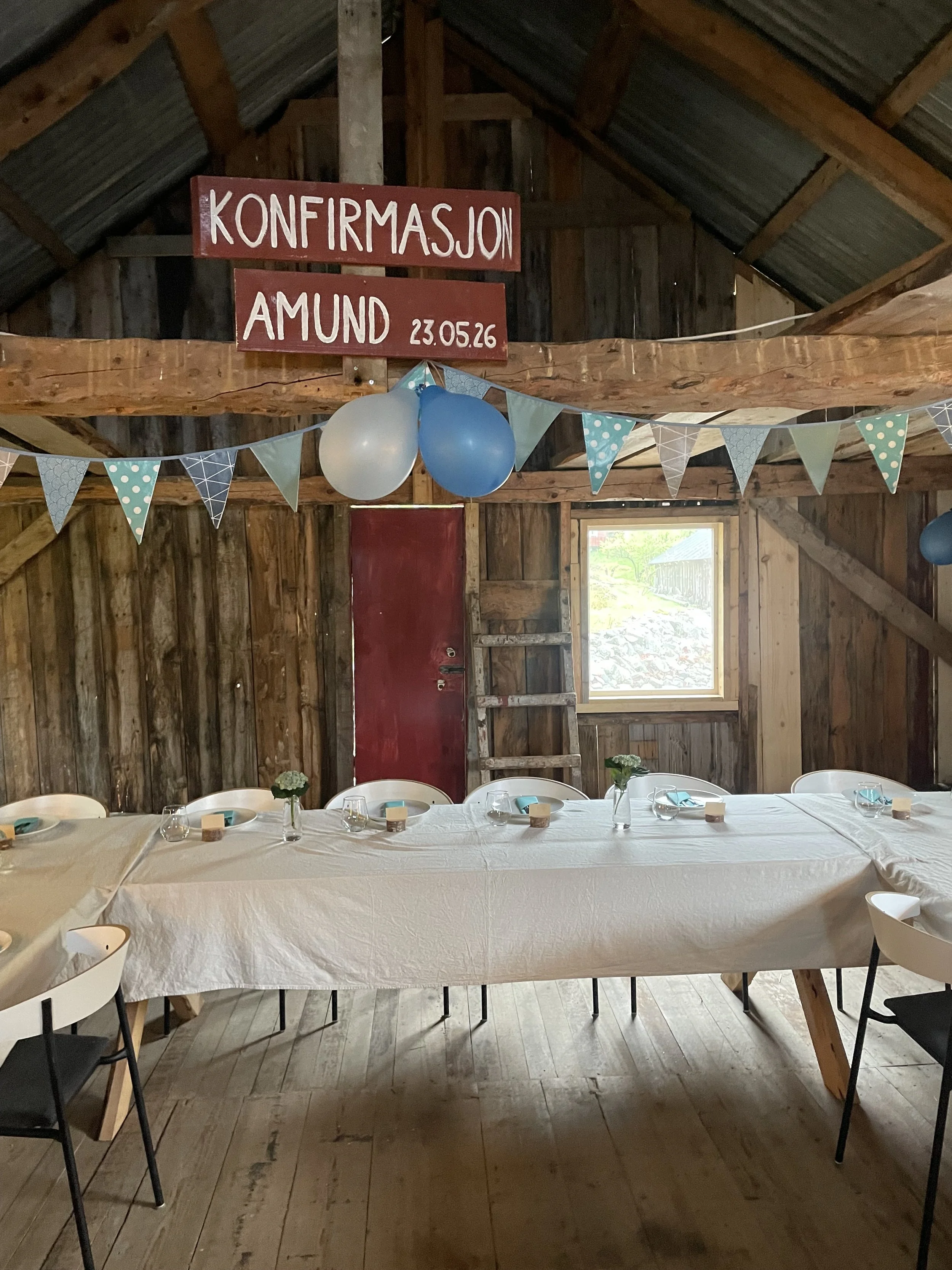 Innendørs bilde av et festbord med hvit duk, små blomster i vaser, tallerkener, glass, og servietter. Bakover er en vegg som er i rå treverk med et lite vindu og dekorative ballonger og flagg som sier 'Konfirmasjon Amund 23.05.26'.