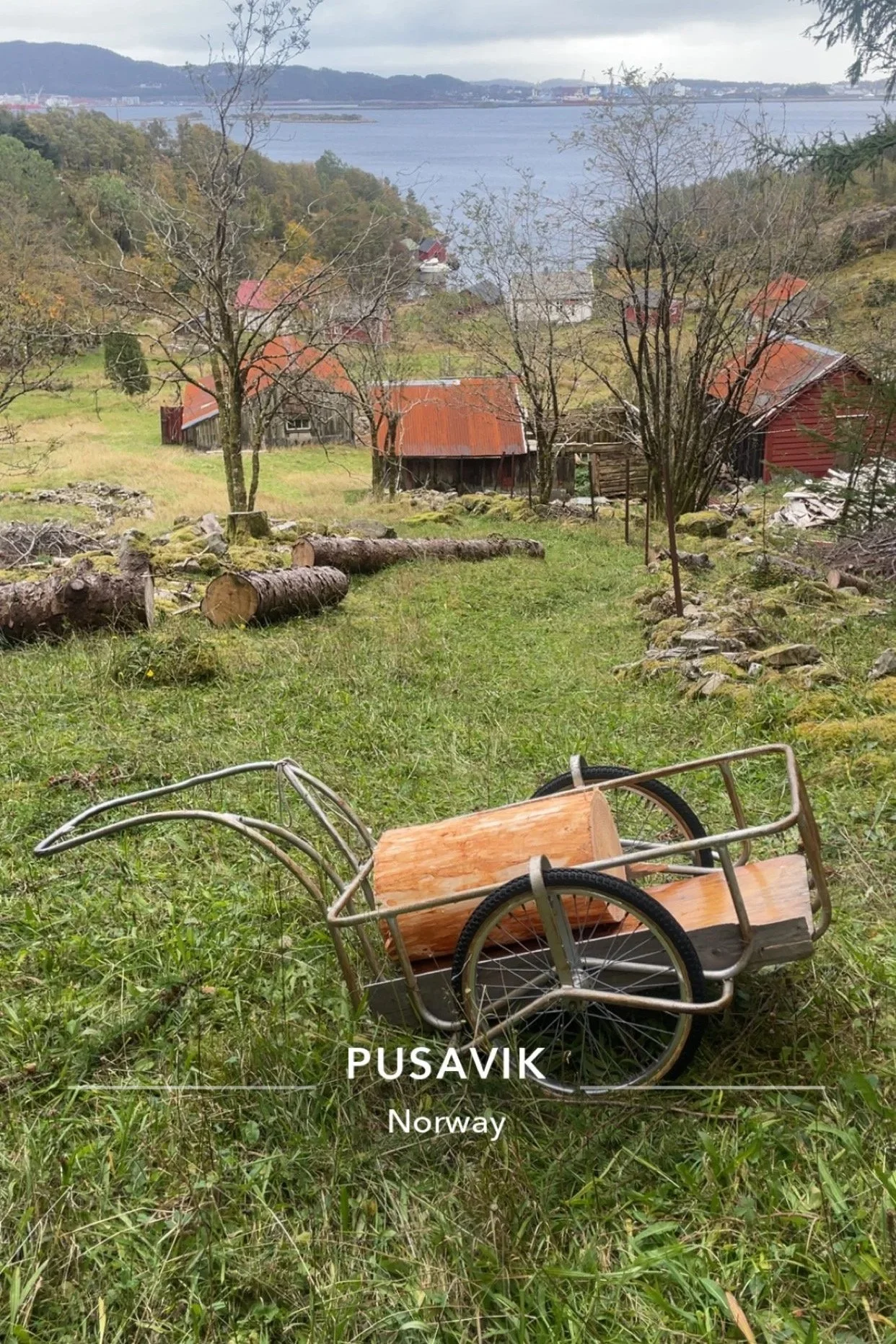 Et utsnitt av et landlig område med trær, noen gamle bygninger med røde tak, og et vann i bakgrunnen under en overskyet himmel. I forgrunnen ligger en trillebår laget av metall med et lite hjul og et trebenees understell, plassert i gresset.