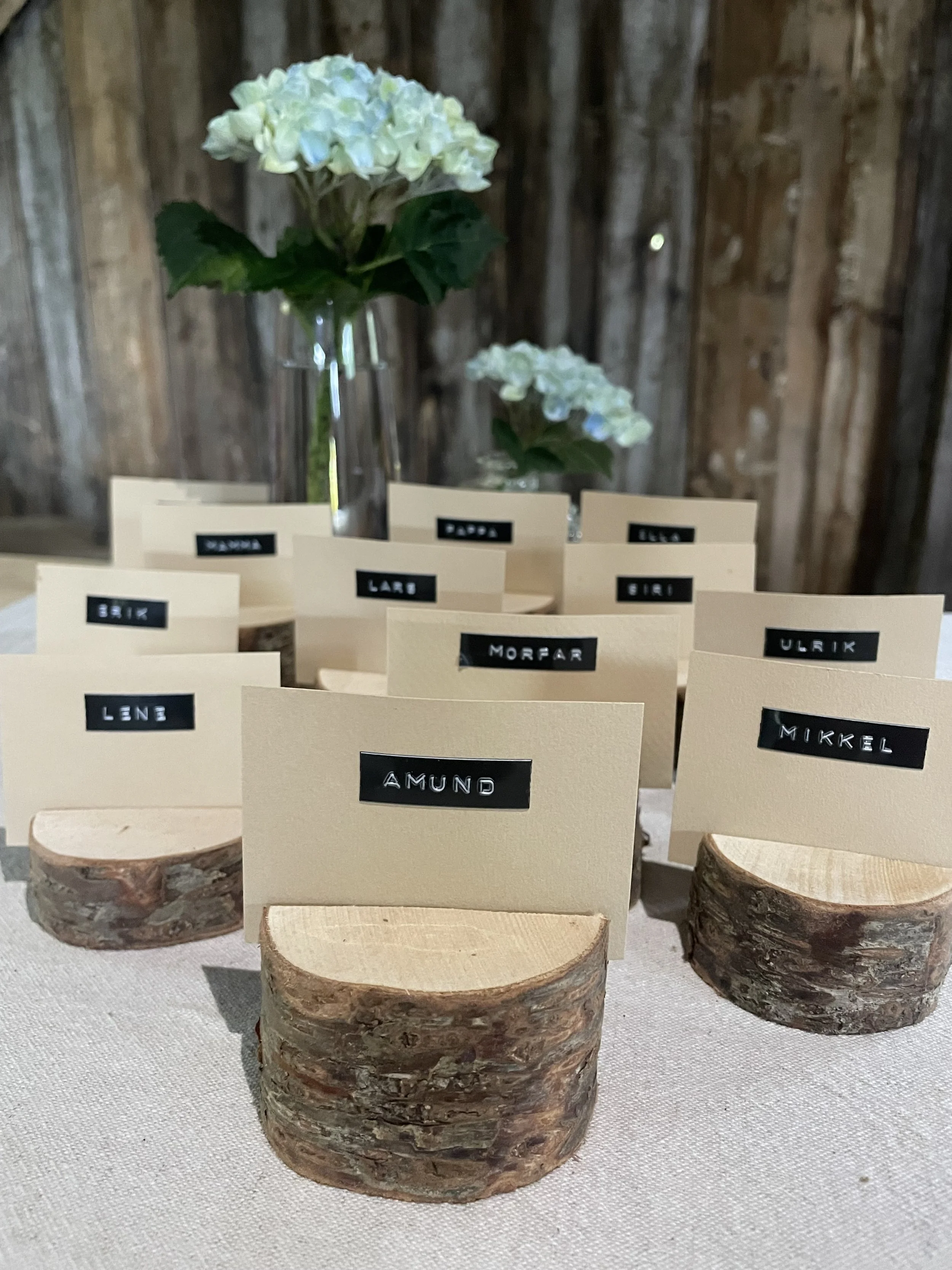 En blomsterbukett med hvite blomster i en glassvase, plassert på en treplate. I forgrunnen er flere små tregavler med navn som 'LENE', 'AMUND', 'MORFAR', 'ULRIK', 'MIKKEL', 'BIRK', 'LARS', 'BODO', og 'ELSE', som sannsynligvis er dekorative bordkort.
