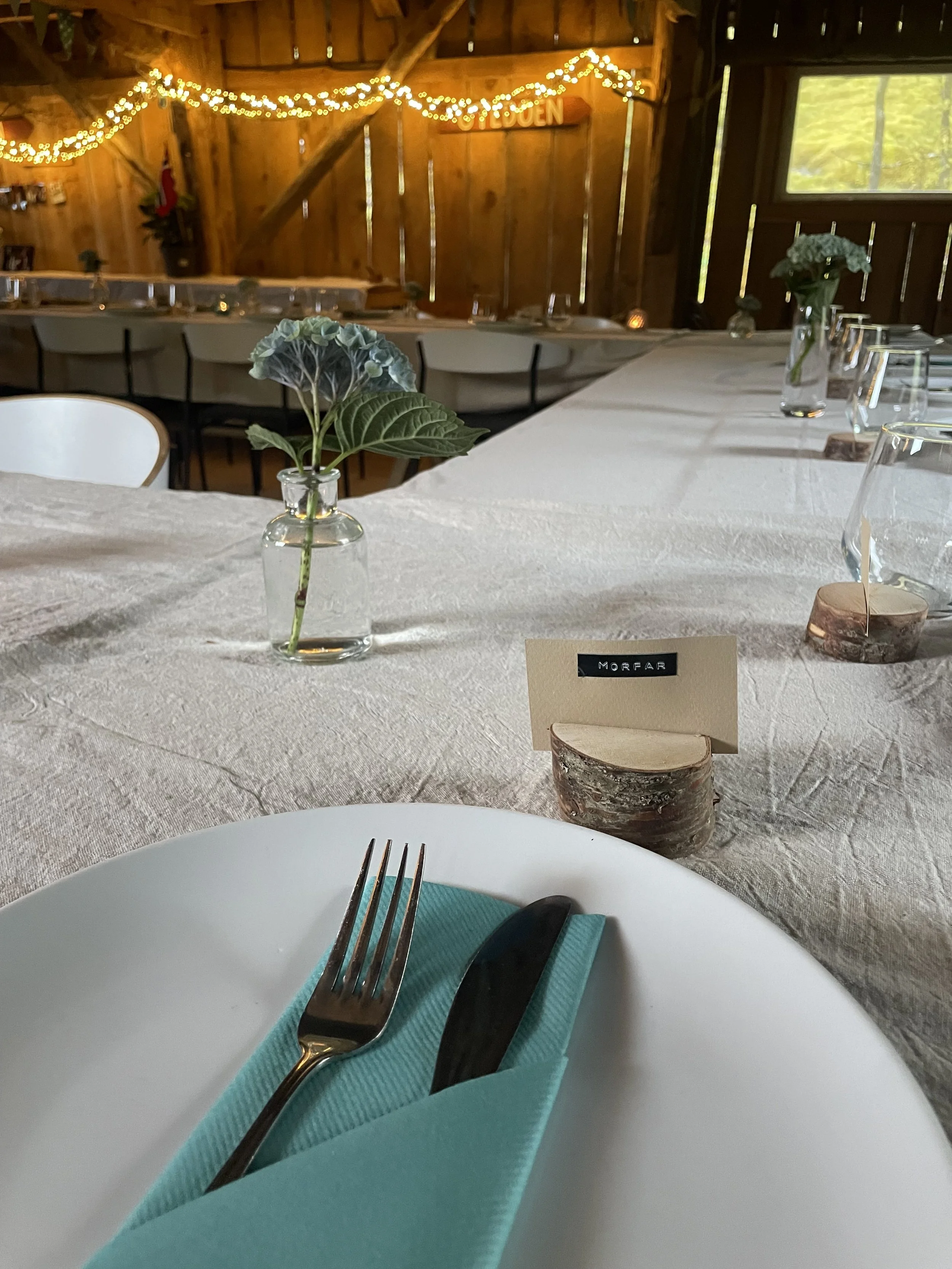 Innendørs spiseområde i et rom med trevegger. Bord er dekket med hvit duk og har små vaser med blomster. En tallizone med en taklampe og julelys dekorerer veggen. Plass for å sette seg til bords med tallerkener, bestikk og glass. Det er en noelstjern