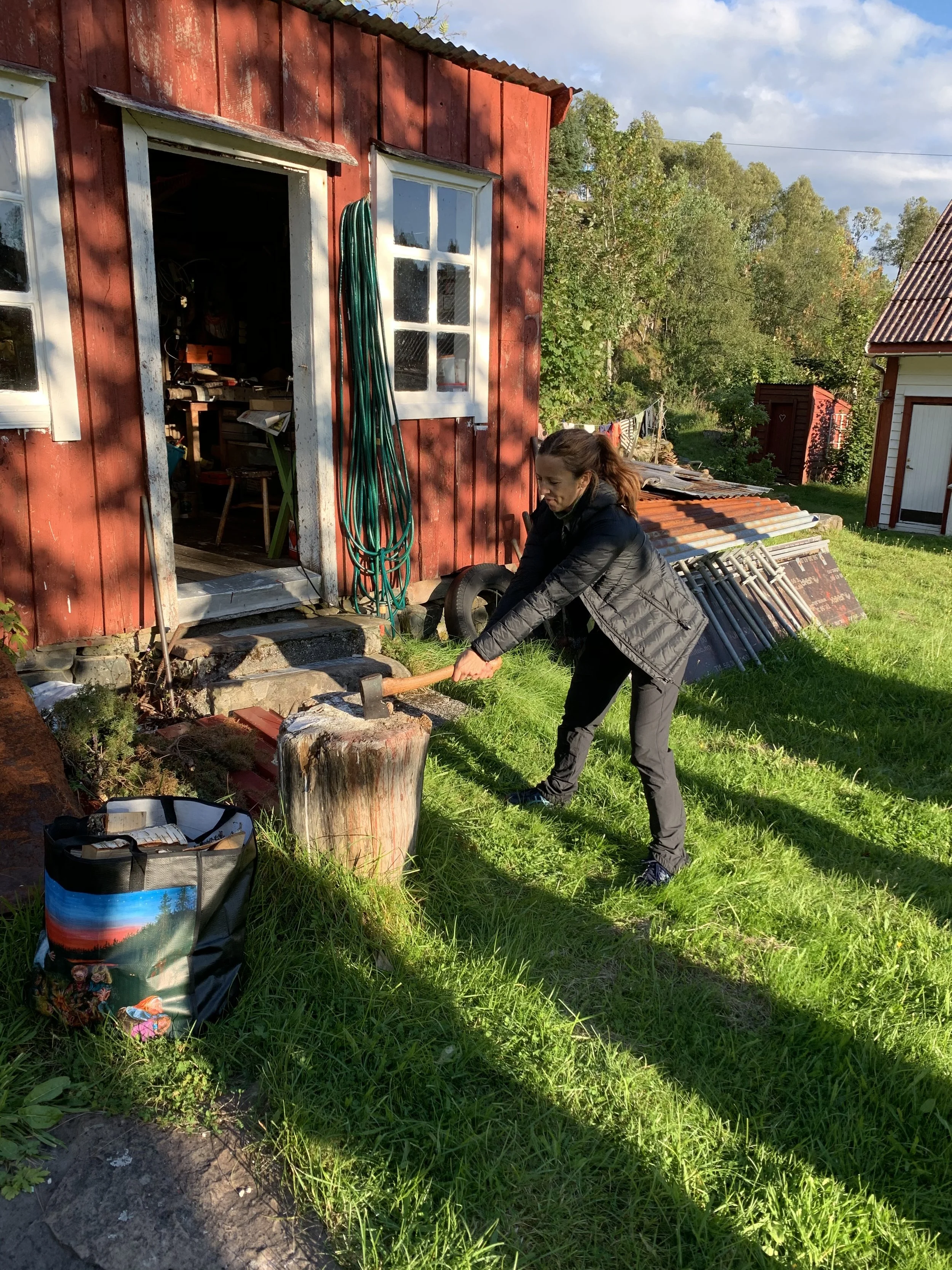 Kvinne som hogger ved med øks foran en rød hytte med hvite vinduer og en hage. Det er sol og grønt landskap i bakgrunnen.