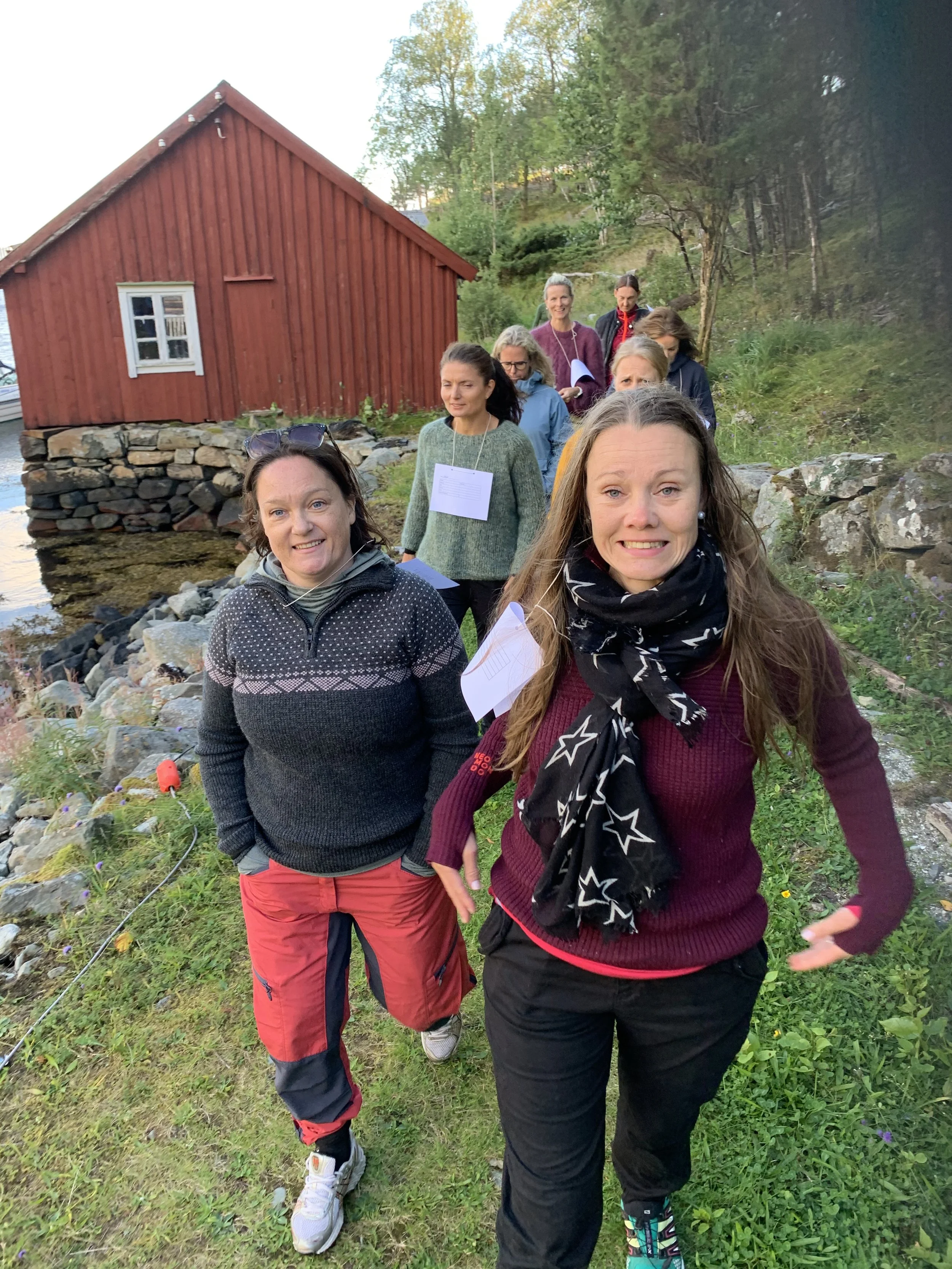 Ti kvinner går langs en sti på landet, med en rød husvegg og grønne trær i bakgrunnen. De har på seg utendørs klær og holder papirer, de ser ut til å delta i en utflukt eller et møte.