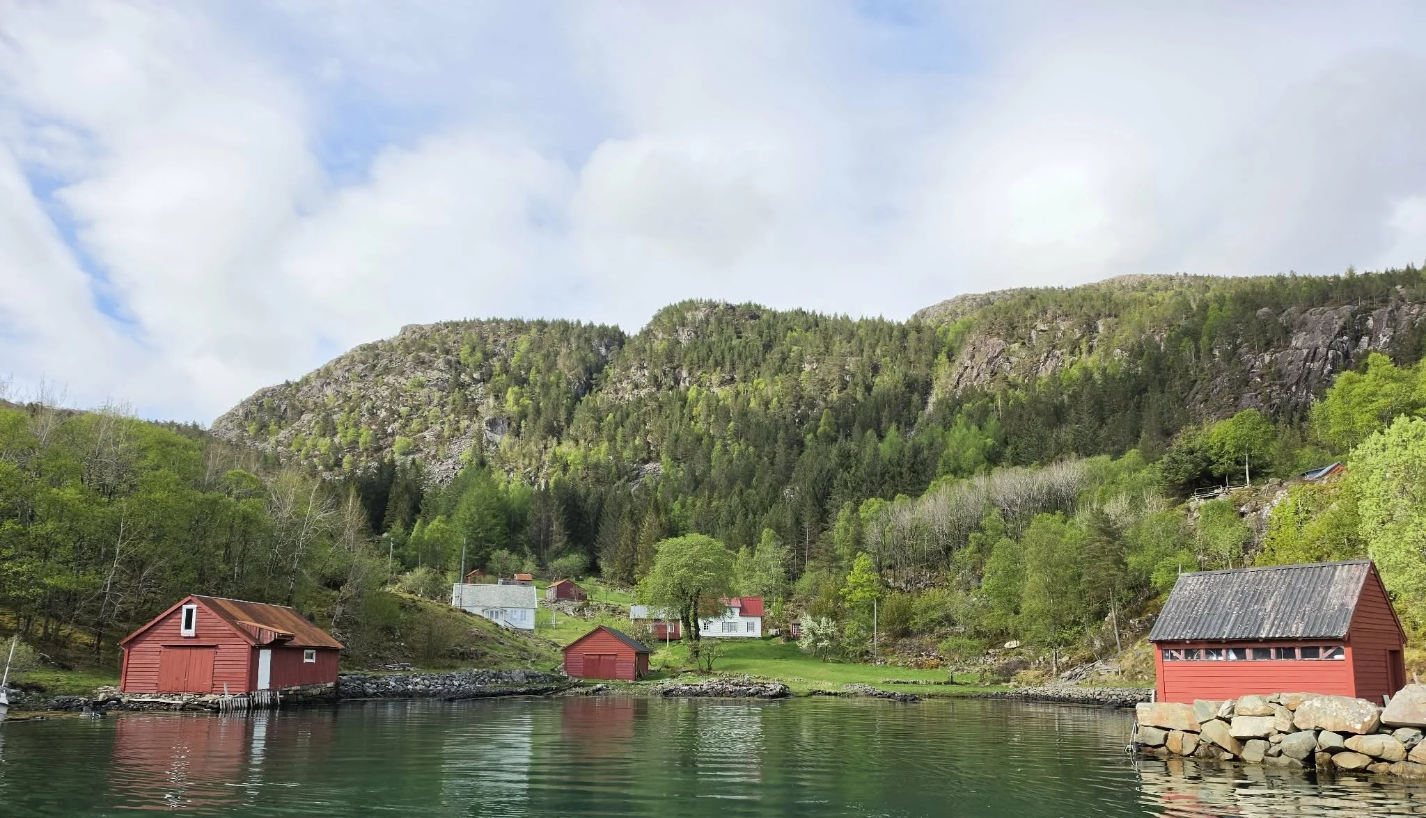 Fjell, grønne trær, små hus ved vann, skyet himmel.
