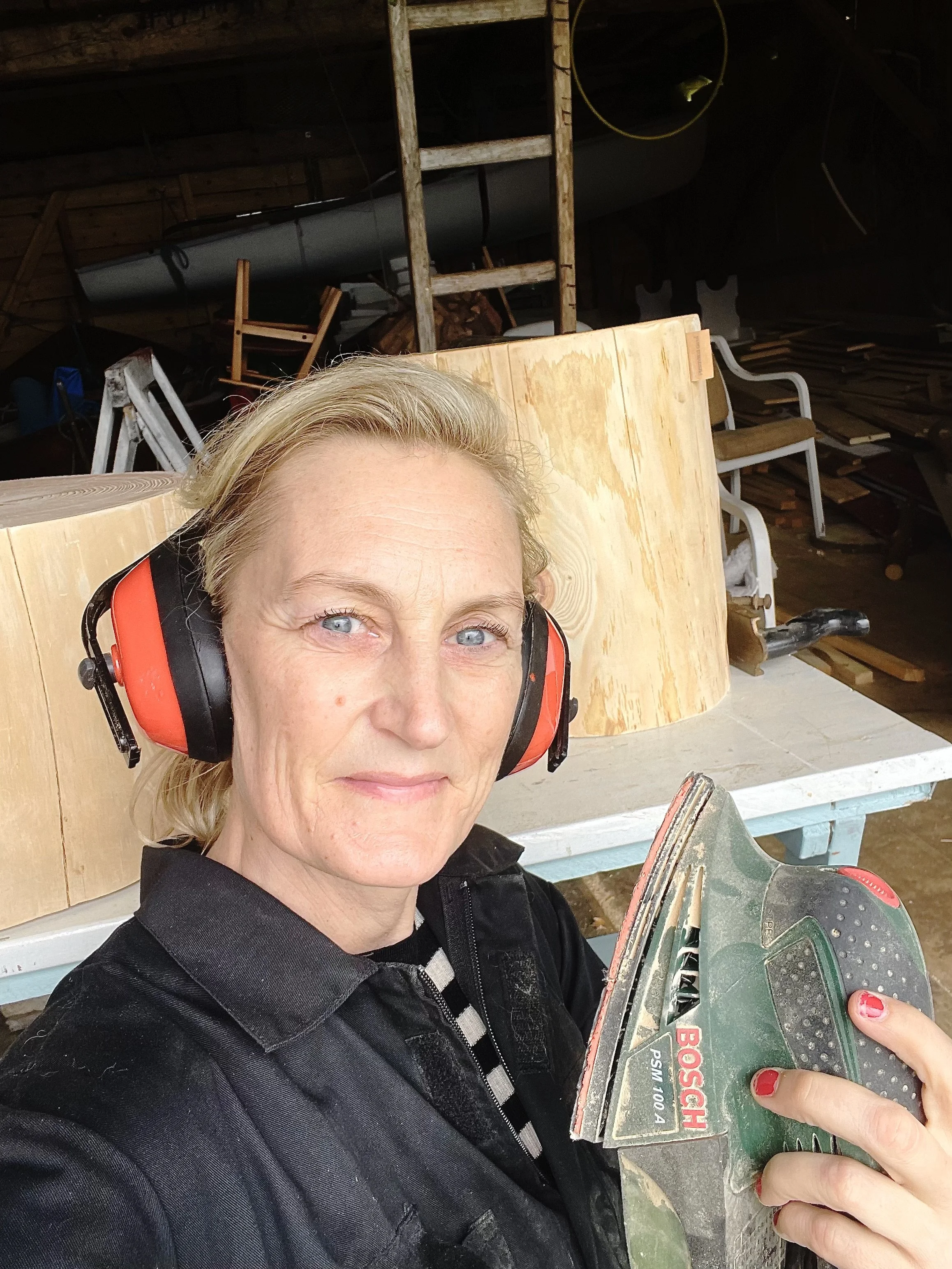 Nærbilde av en kvinne med lyseblå øyne og blondt hår, iført verneutstyr og svart arbeidsjakke, som holder en Bosch-nerbesliper, i et verksted eller skur med trestabler, verktøy og trebiter i bakgrunnen.