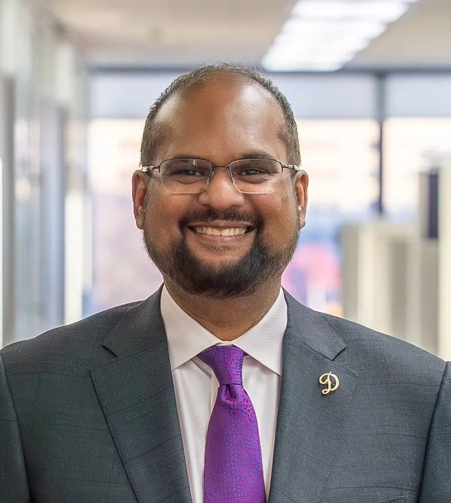 A smiling man in a gray suit, white shirt, and purple tie, wearing glasses and a lapel pin, in an office setting.
