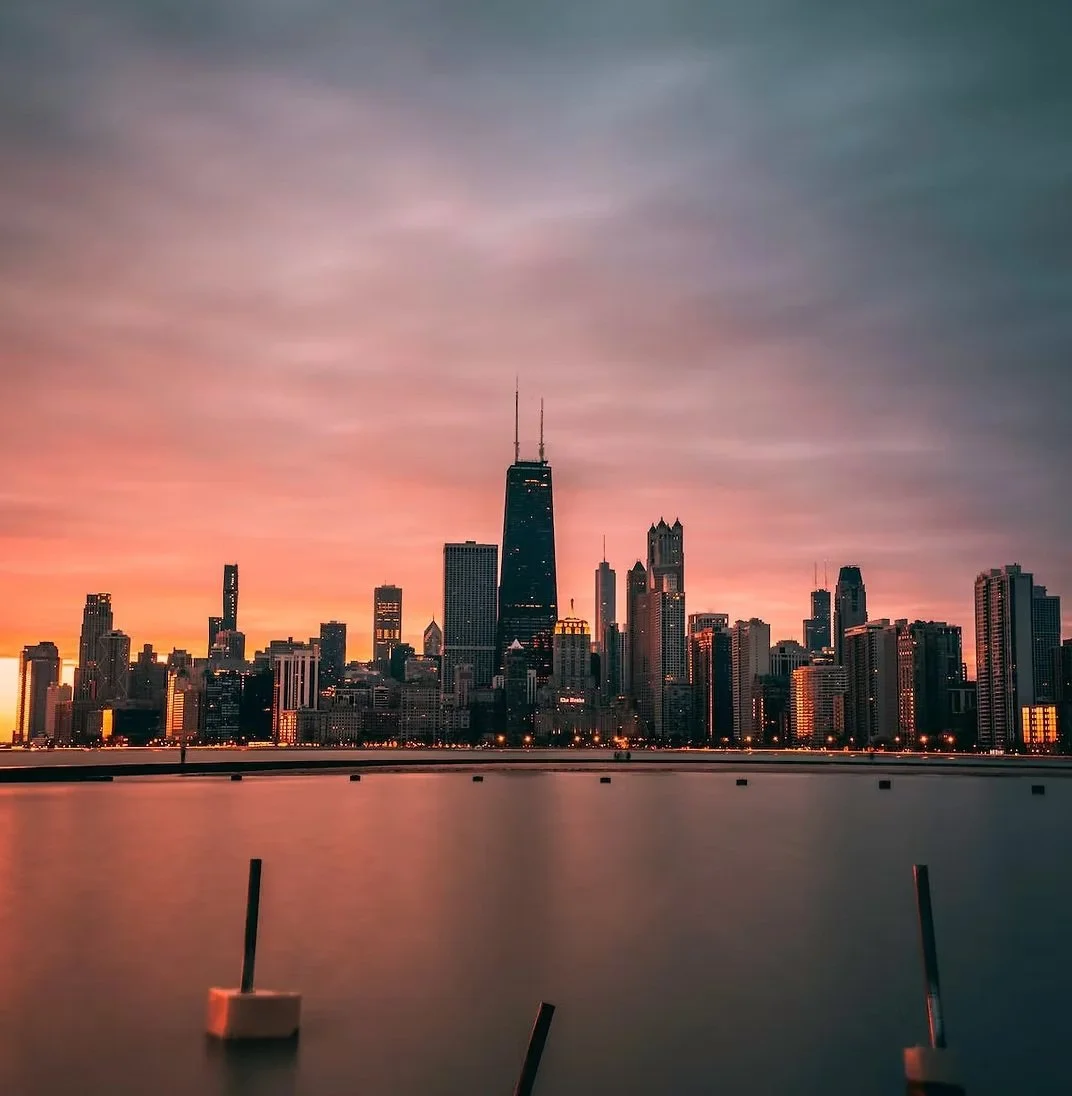 Chicago skyline at sunset with the Willis Tower prominently visible, reflected in calm water with boats and docks in the foreground.
