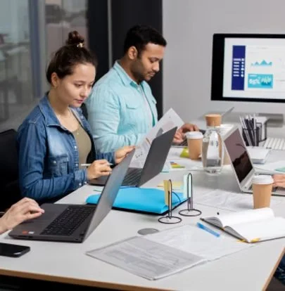 Two people working at a desk with laptops and documents in an office, analyzing data on a computer screen.