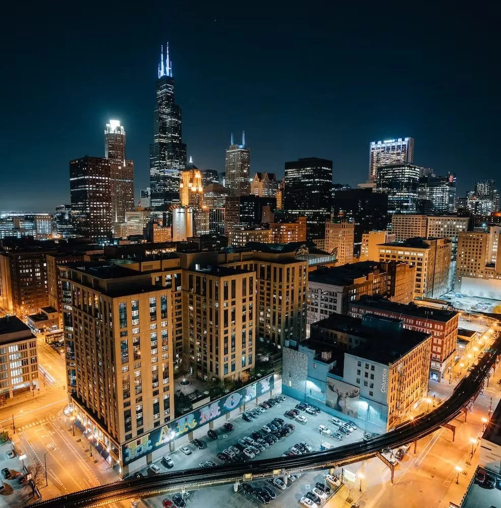 Nighttime aerial view of downtown Chicago featuring the tall Willis Tower with blue lights on top, surrounded by other illuminated skyscrapers and city buildings, with streets and parking lots below.