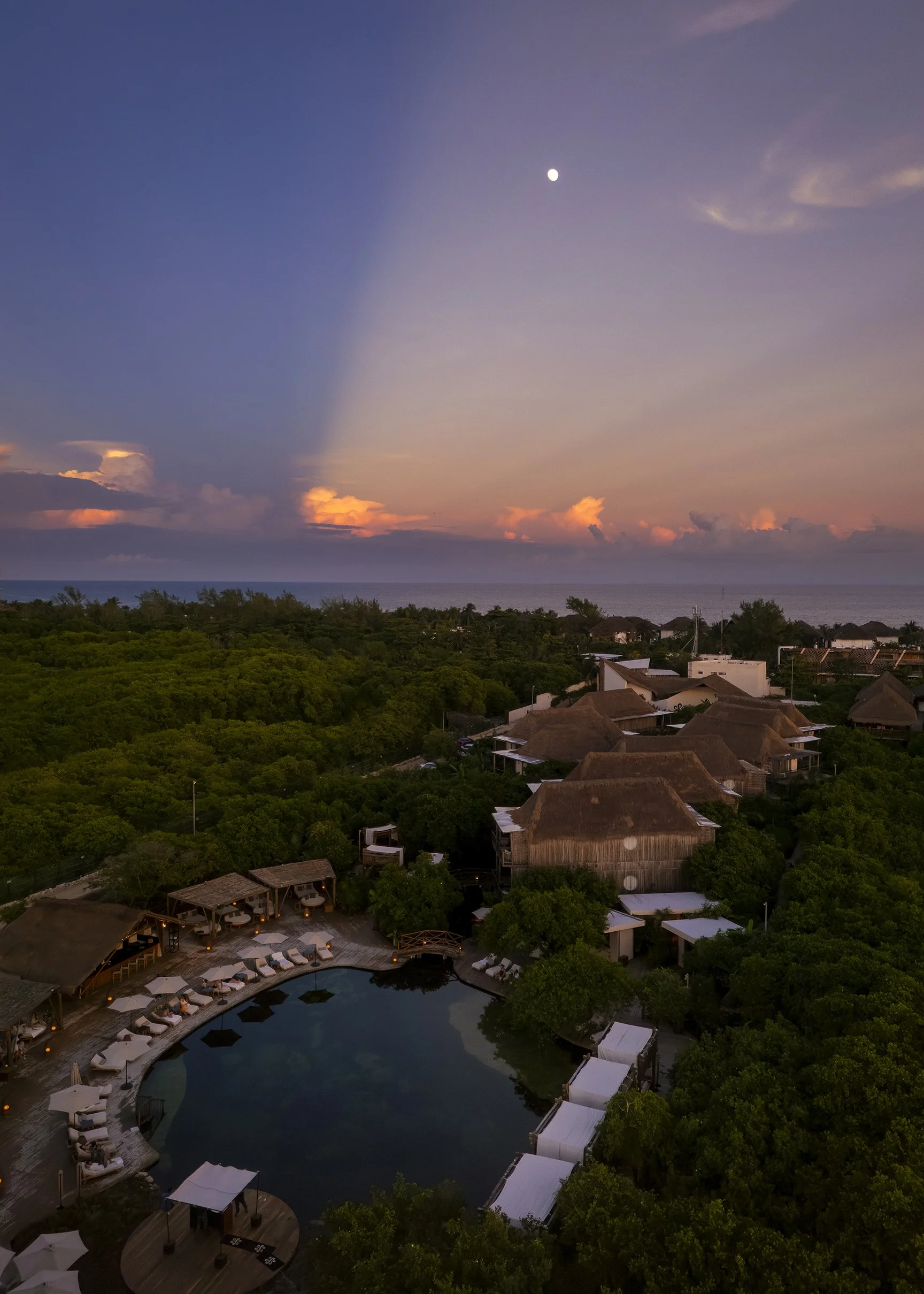 A tropical resort at sunset featuring a swimming pool, thatched-roof buildings, lush green trees, and a view of the ocean in the distance, with a partly cloudy sky and visible moon.