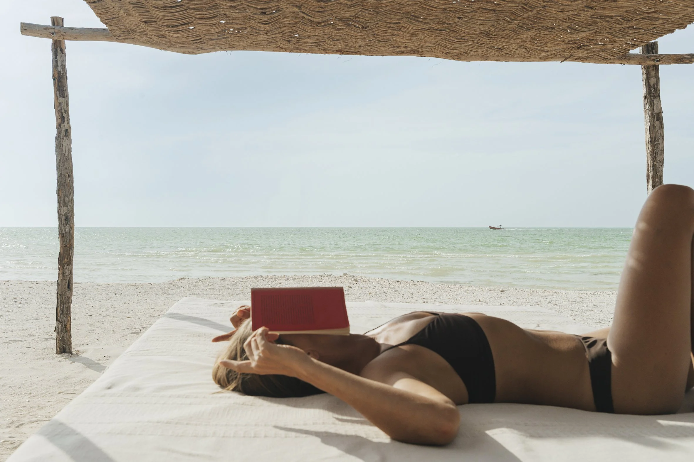 A person lying on a beach lounge under a thatched umbrella, reading a red book, with the ocean in the background and a boat on the water.