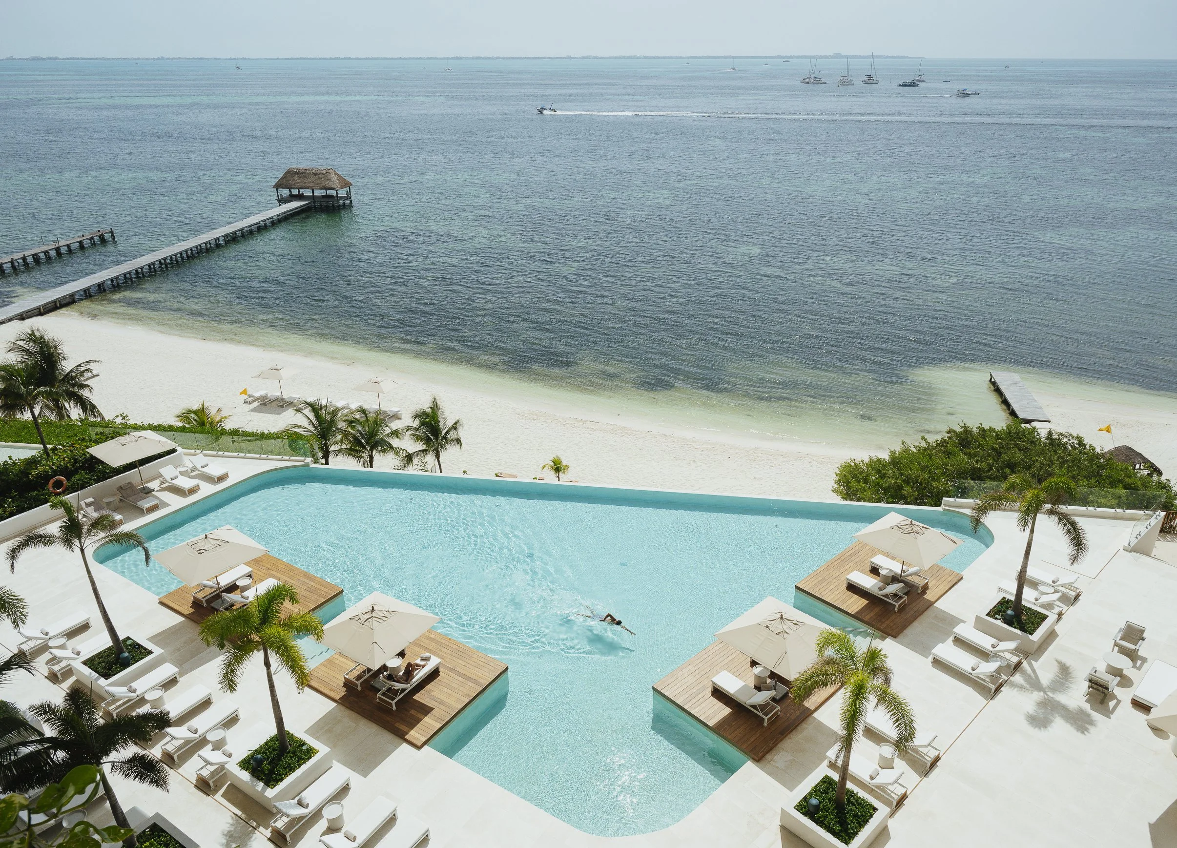 View of a seaside resort in cancun with a swimming pool surrounded by lounge chairs and umbrellas, a dock with a thatched-roof gazebo extending into the ocean, and several sailboats and boats on the water in the background.