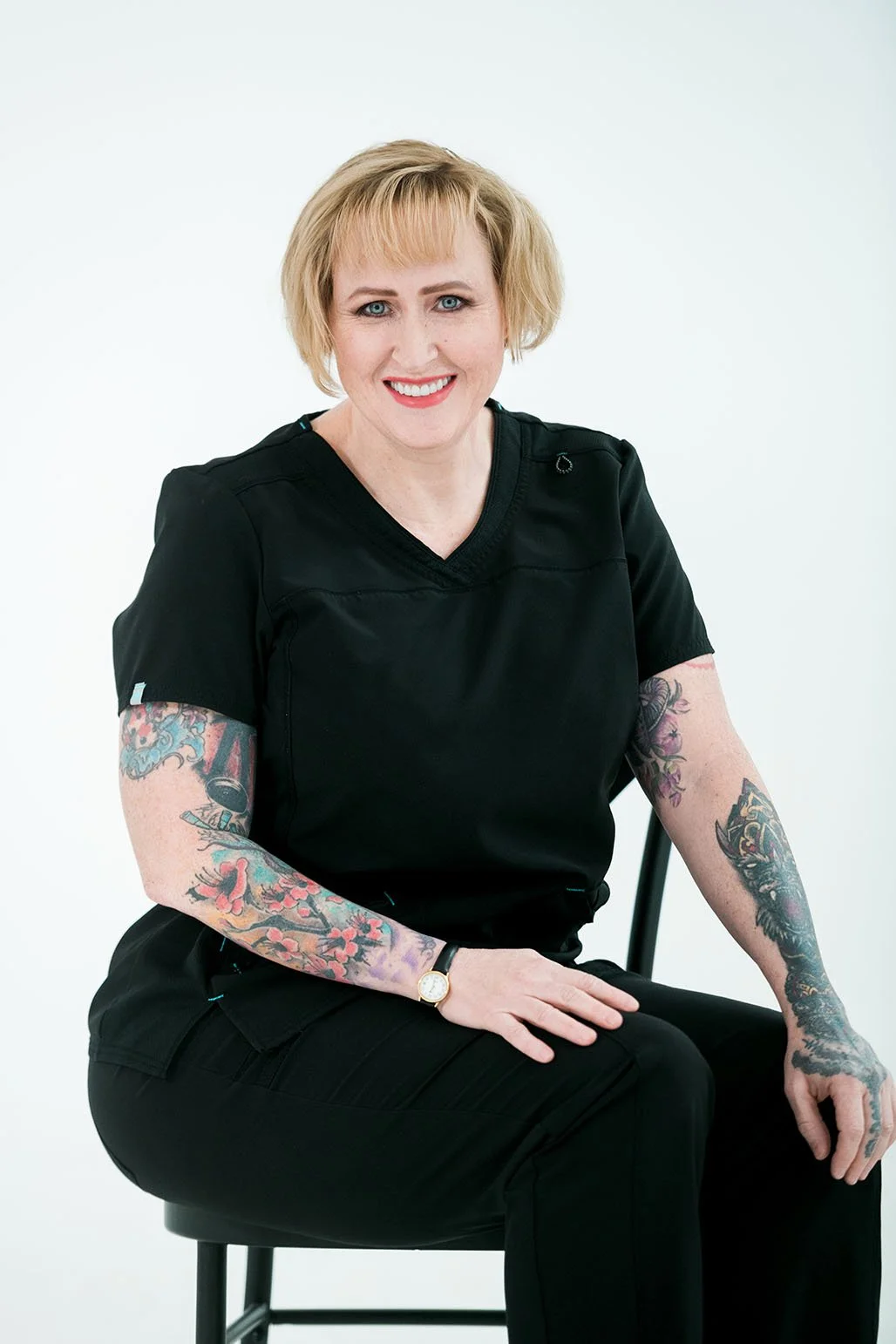 A woman with blonde hair, tattoos on her arms, wearing black scrubs, sitting on a chair, smiling, against a plain white background.