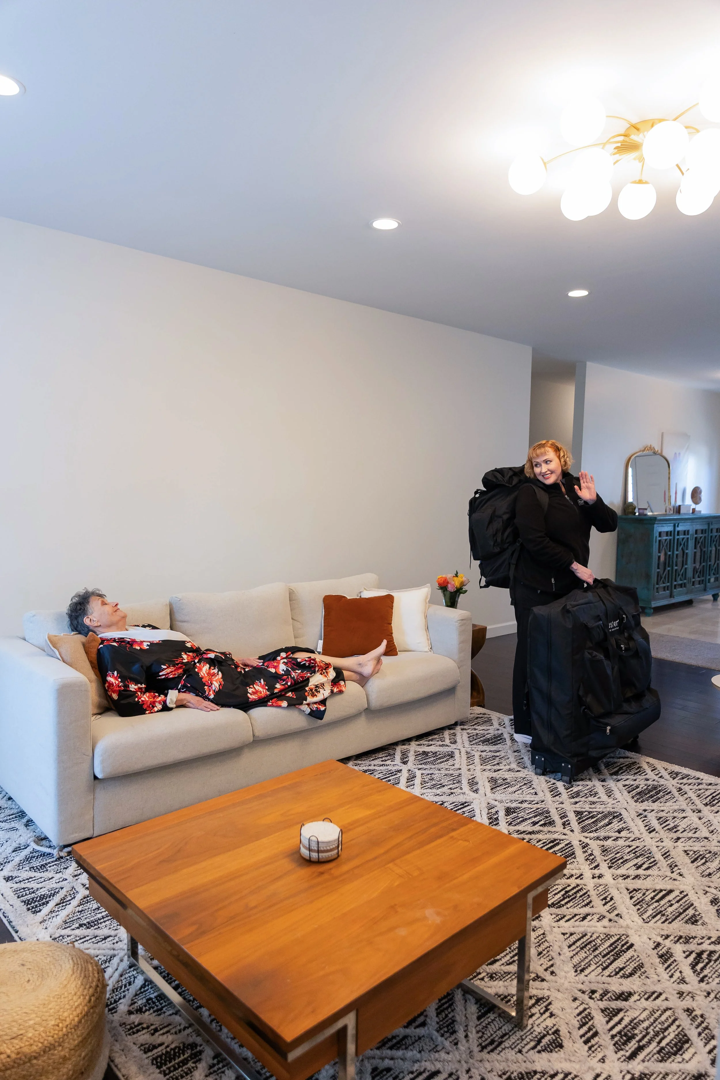 A woman in a black jacket and backpack greeting a woman lying on a beige sofa in a living room. The woman on the sofa is wearing a black dress with a floral pattern and appears to be resting. There is a rectangular wooden coffee table with a small decorative item on it, a patterned rug, and a blue cabinet in the background. The living room has white walls and ceiling, with a modern chandelier hanging from the ceiling.