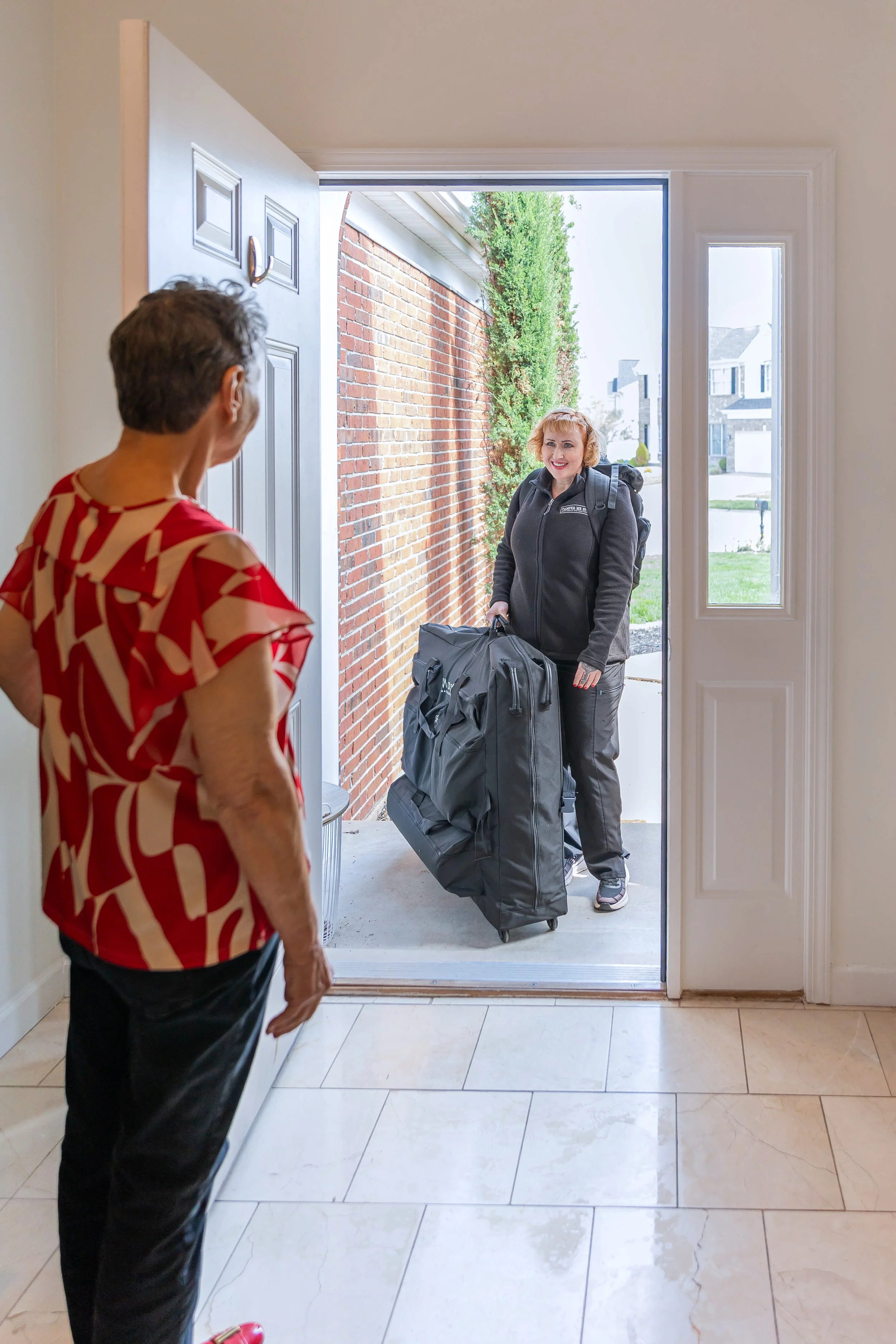 A woman with a red and white patterned top standing inside near the doorway, talking to a woman outside with a black jacket and backpack who is arriving with a large rolling bag.
