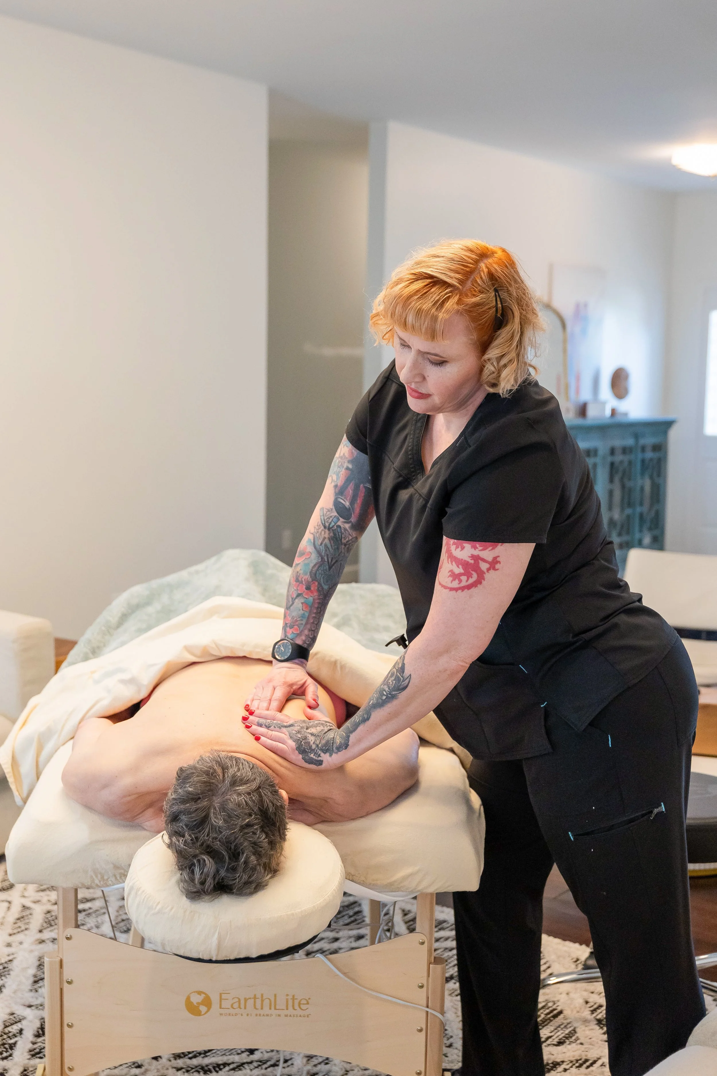 A woman with tattoos giving a massage to a person lying face down on a massage table in a well-lit room.