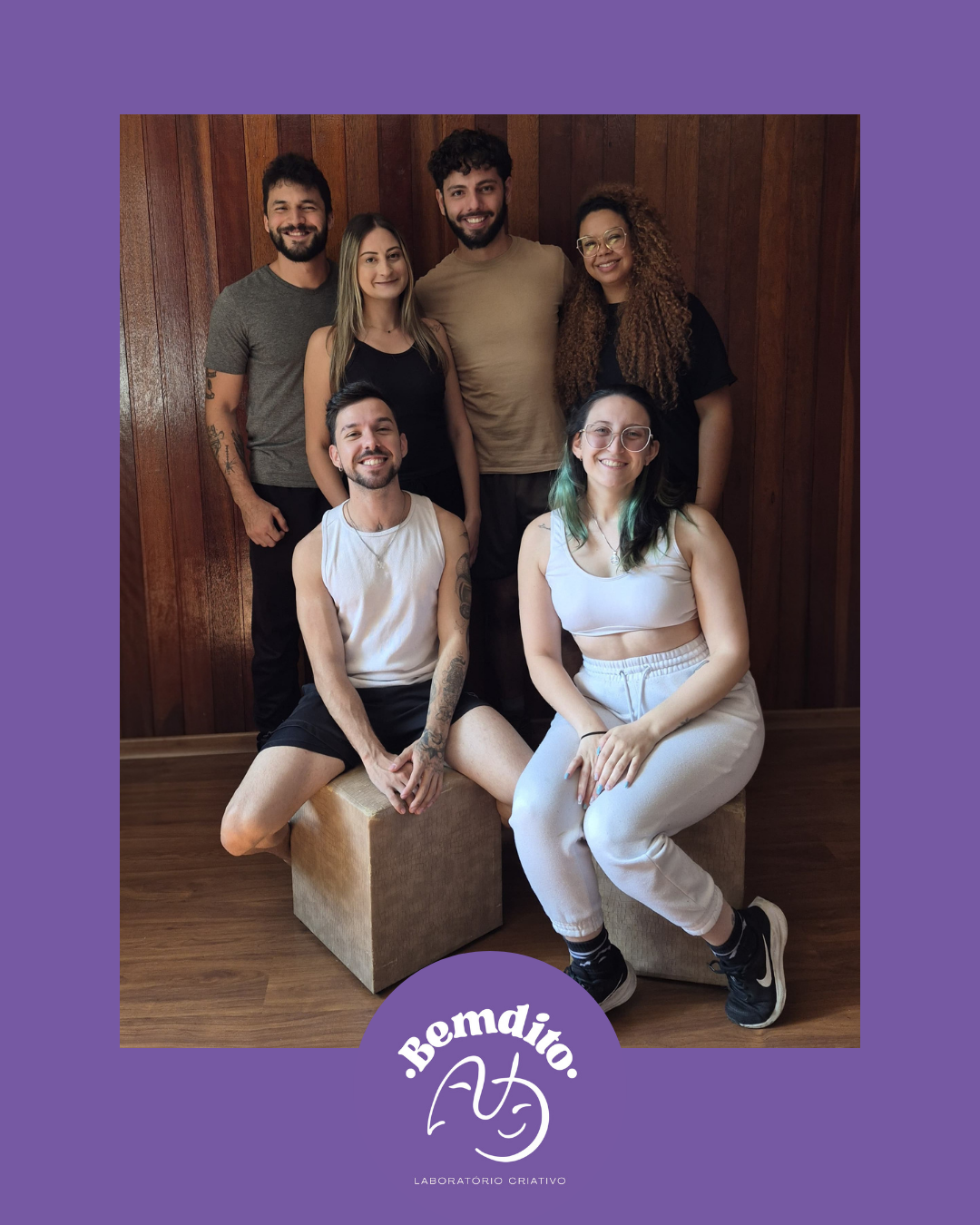 Grupo de sete pessoas sorrindo, posando na frente de uma parede de madeira. Duas pessoas estão sentadas em blocos de cortiça, enquanto as outras cinco estão de pé. Todos parecem felizes e descontraídos. Logo na parte inferior da imagem, há uma marca d'água com o texto 'Bemdito' e um desenho de rosto sorridente, além da frase 'Laboratório Criativo'.