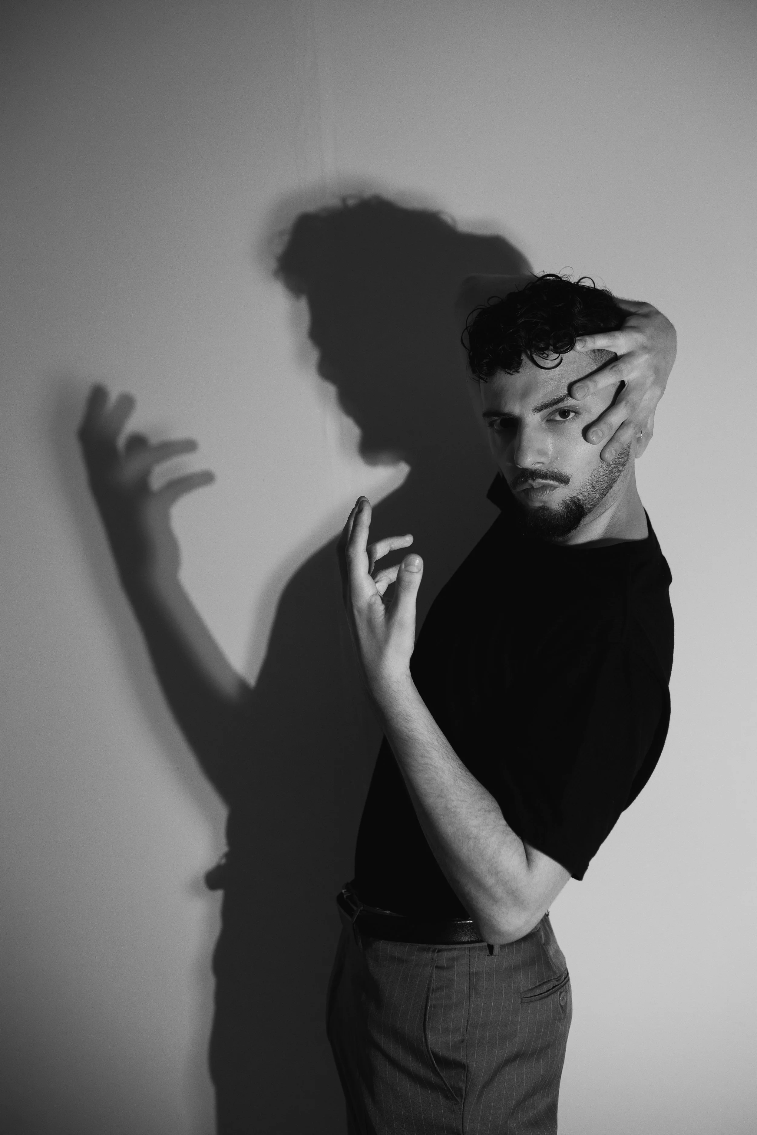Homem com cabelo cacheado segurando a cabeça com uma mão e a outra mão na altura do rosto, ao lado de uma parede, criando uma sombra de sua mão na parede.