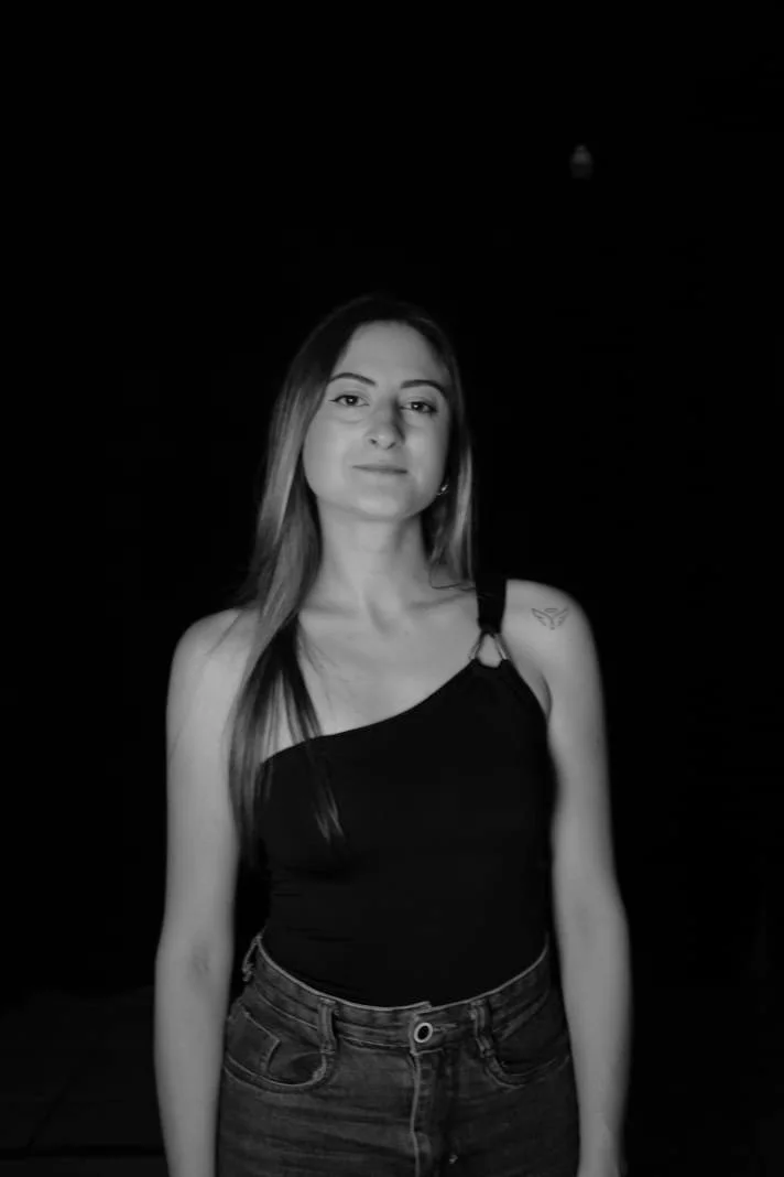 Jovem mulher com cabelo liso e olhos claros, vestindo regata preta e jeans, com tatuagem no ombro esquerdo, posando contra fundo escuro.