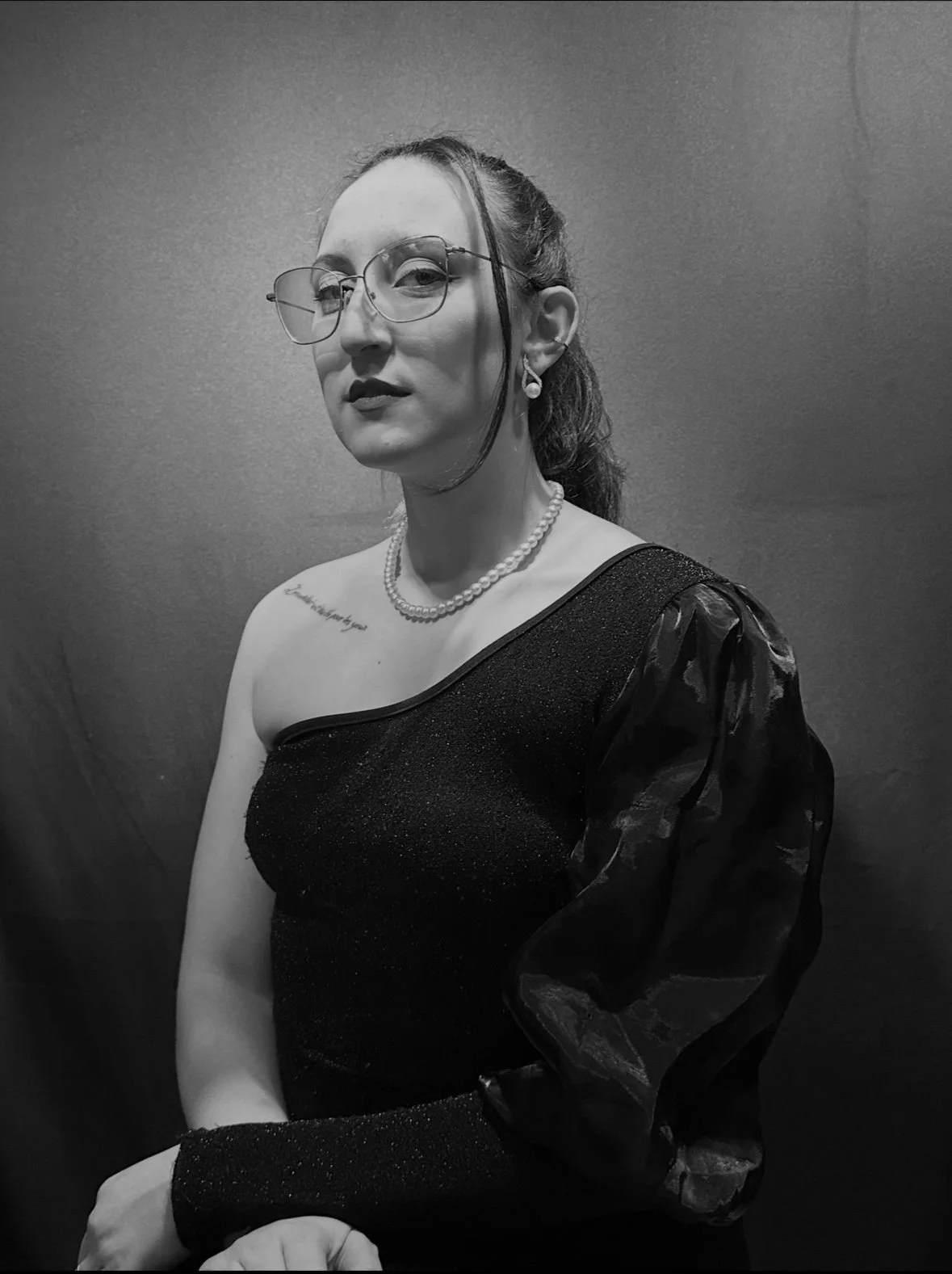 Retrato em preto e branco de uma mulher vestida com vestido negro, usando óculos, brincos de pérola e colar de pérola, com cabelo preso e expressão séria, contra fundo escuro.