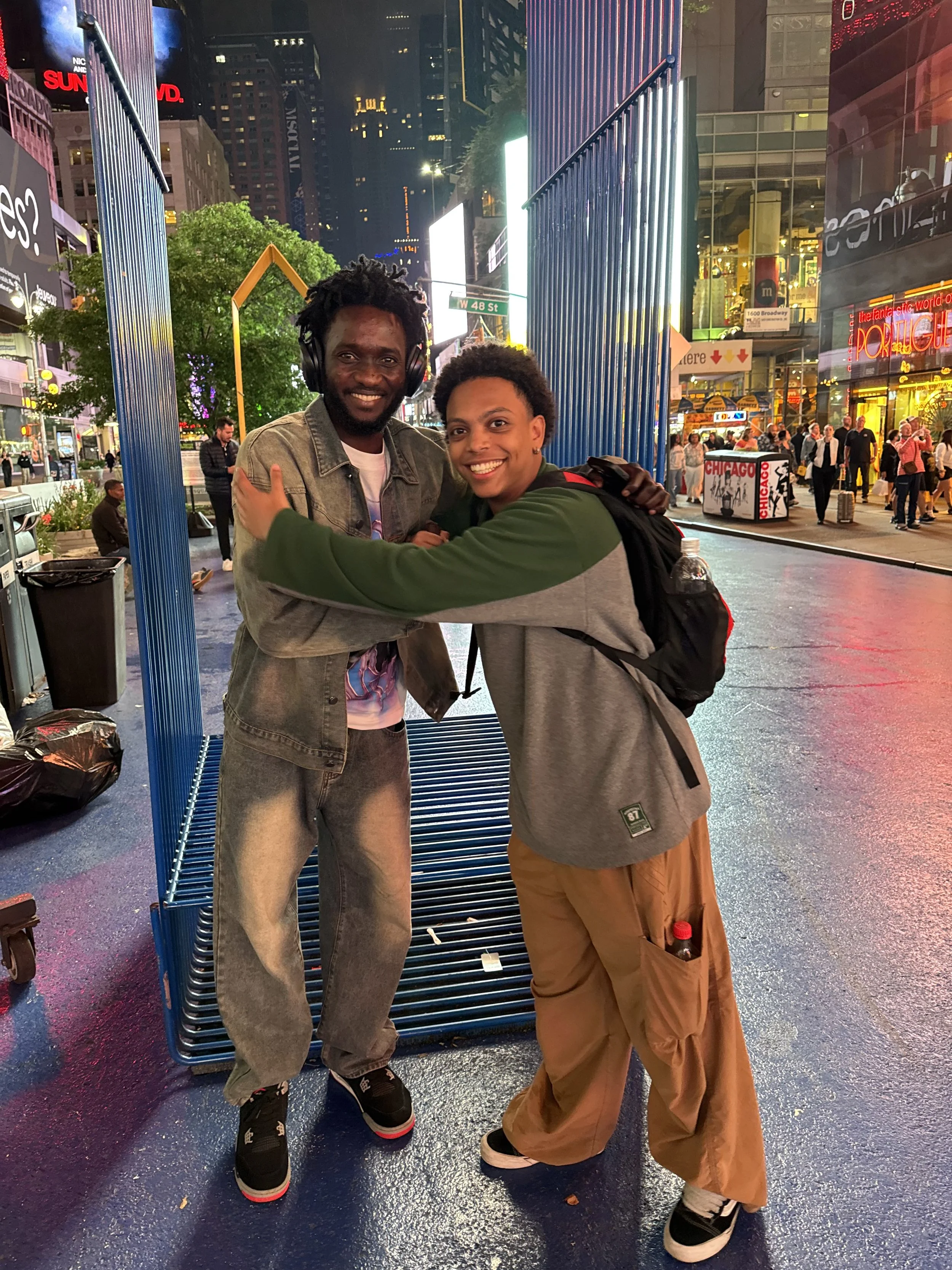 Two young men smiling and hugging each other on a busy New York City street at night, with tall buildings, digital billboards, and illuminated storefronts in the background.