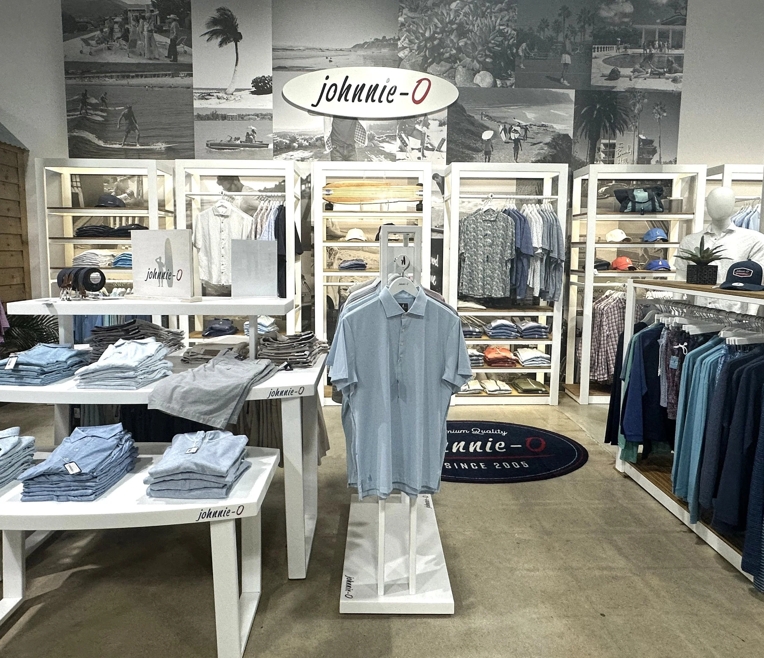 Display inside a retail store showing white shelves with various clothing items, hats, and accessories. A large black oval sign reads 'johnnie-O', and the background features a collage of black and white beach scenes and palm trees.