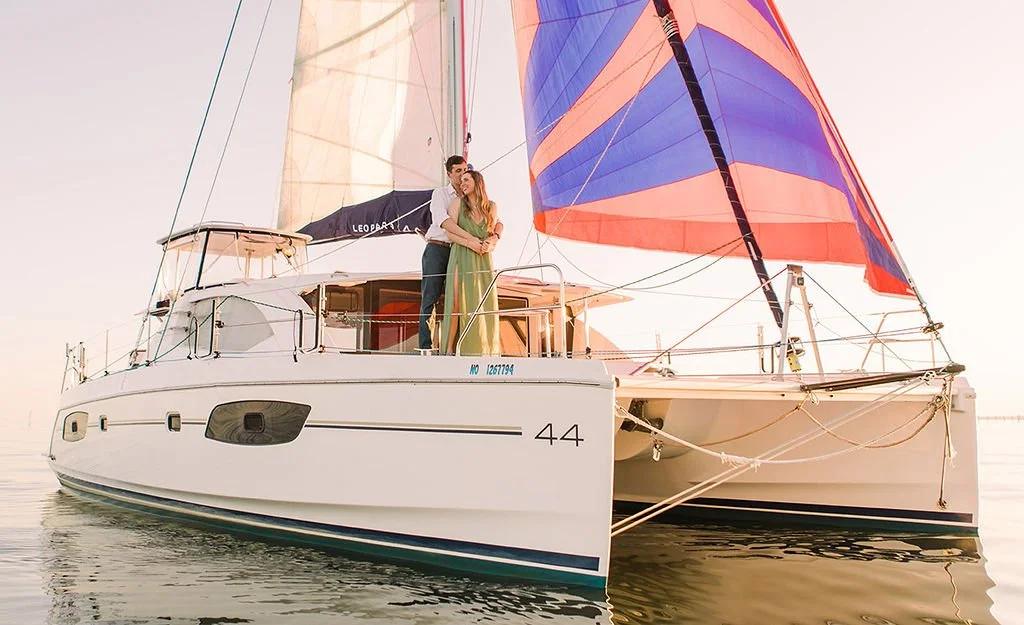 A couple standing on the deck of a sailboat with colorful sails during sunset.