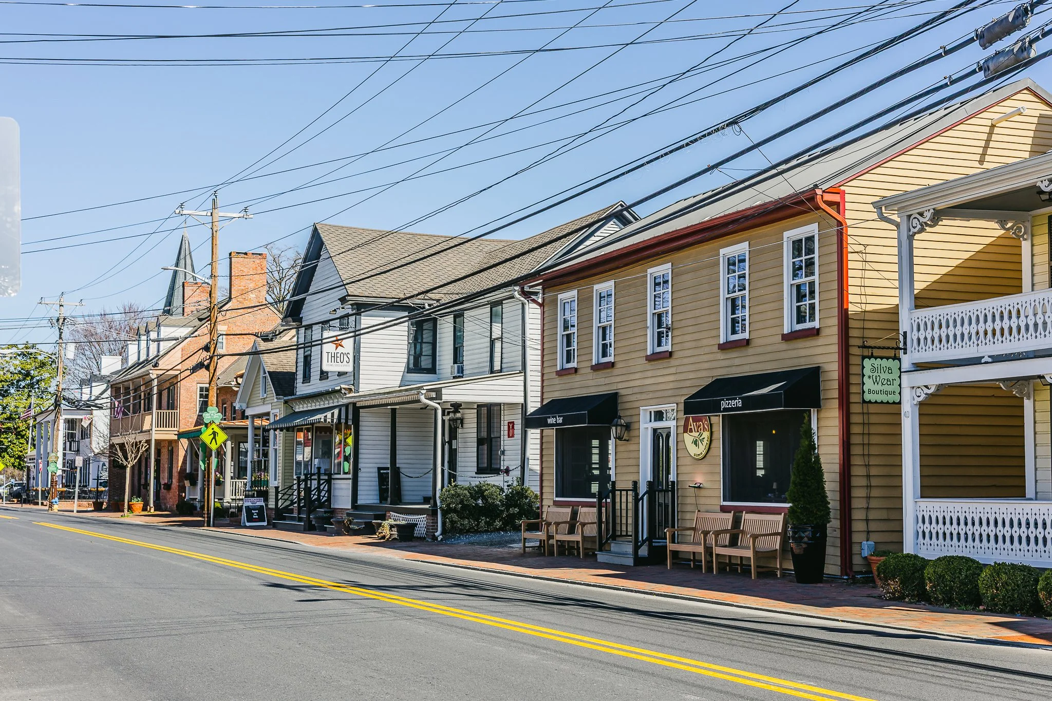 A quaint street with colorful storefronts and residential houses, featuring signage for businesses like a pizzeria and wine bar, and benches on the sidewalk. Power lines cross the clear blue sky above.