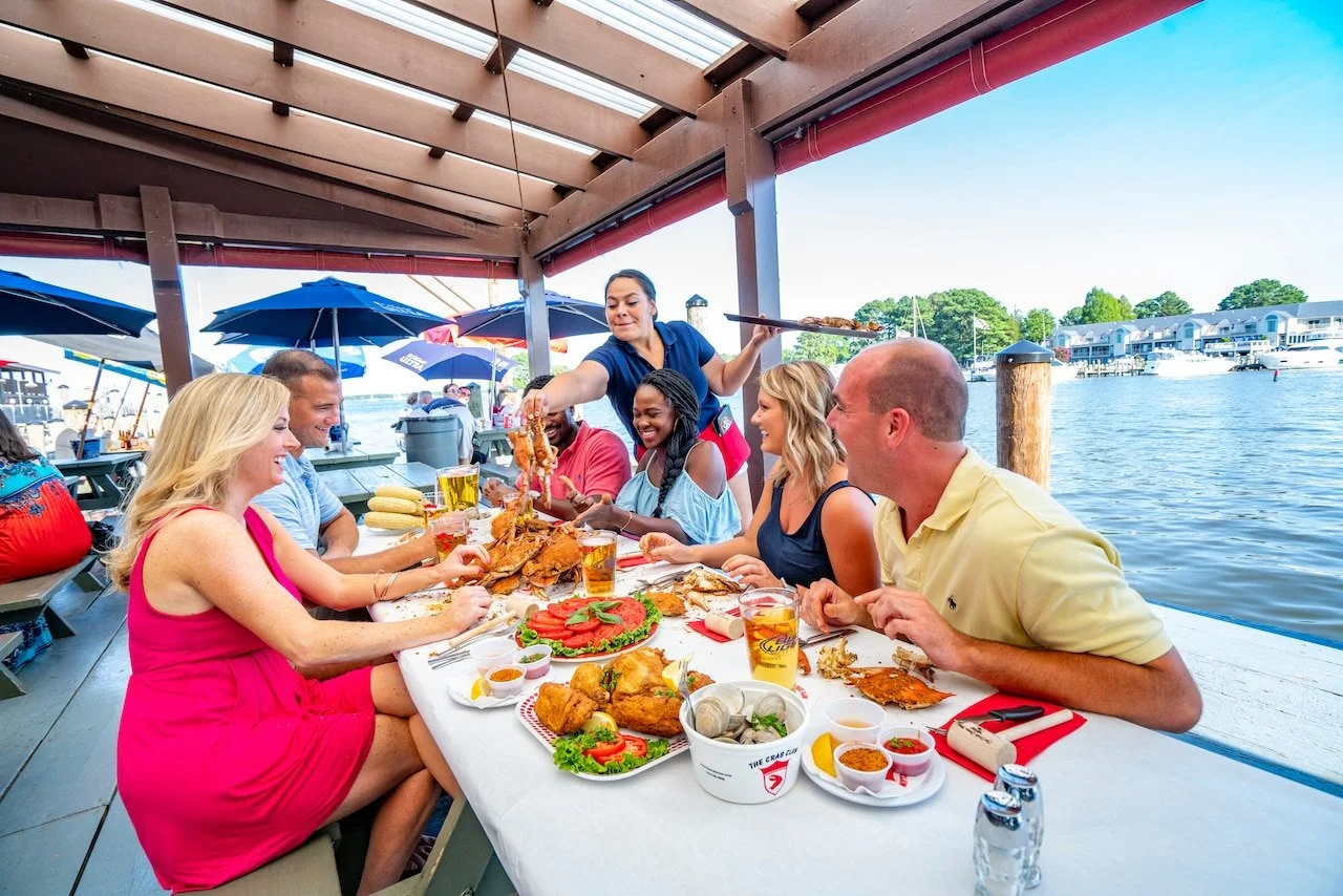 Group of friends dining outdoors by a lake, enjoying seafood and drinks under a covered patio with umbrellas and boats in the background.