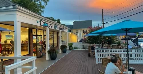 Outdoor patio dining area with customers seated under blue umbrellas, a Comar's store with large windows, and sunset sky in the background.