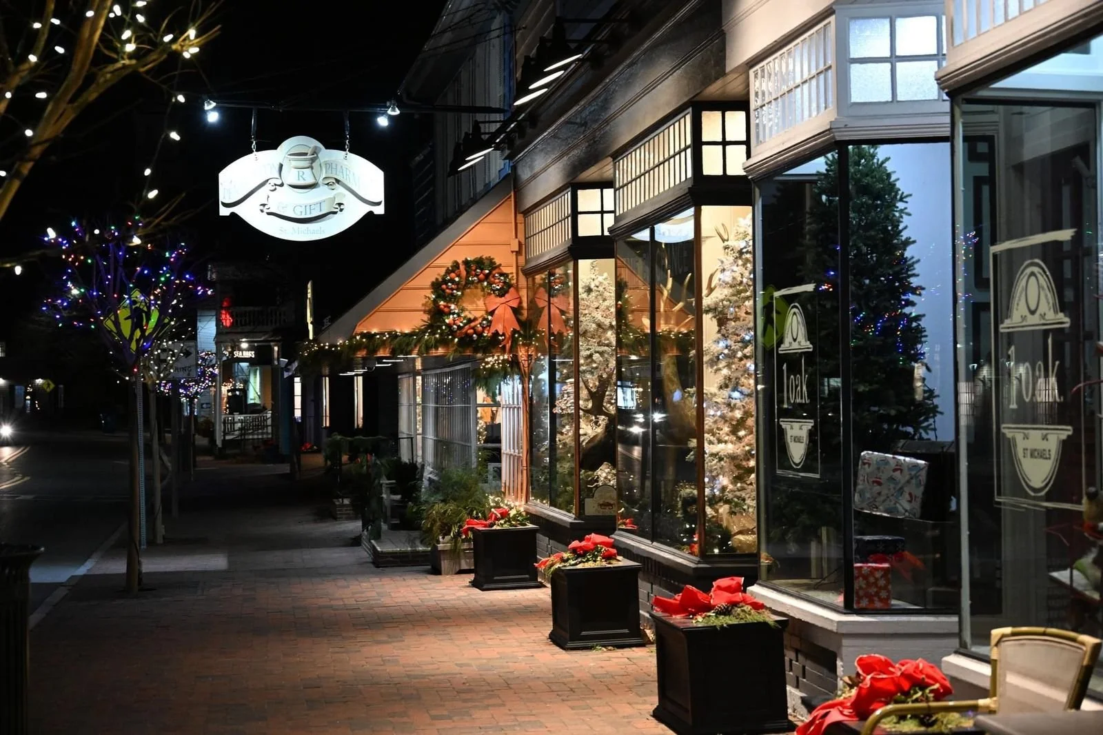 A storefront decorated for Christmas with a Christmas tree, festive wreaths, and red poinsettias outside. The store has large glass windows and is illuminated at night.