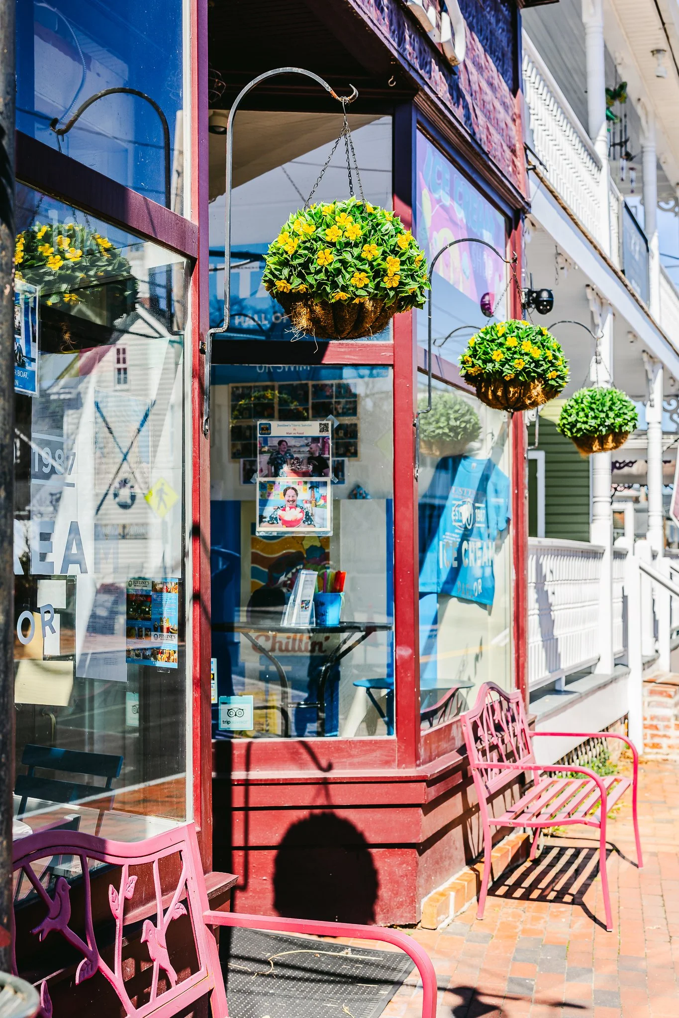 View of a storefront with hanging flower pots containing yellow flowers, pink benches outside, and chalkboard signs, with a reflection of the street and buildings in the glass window.