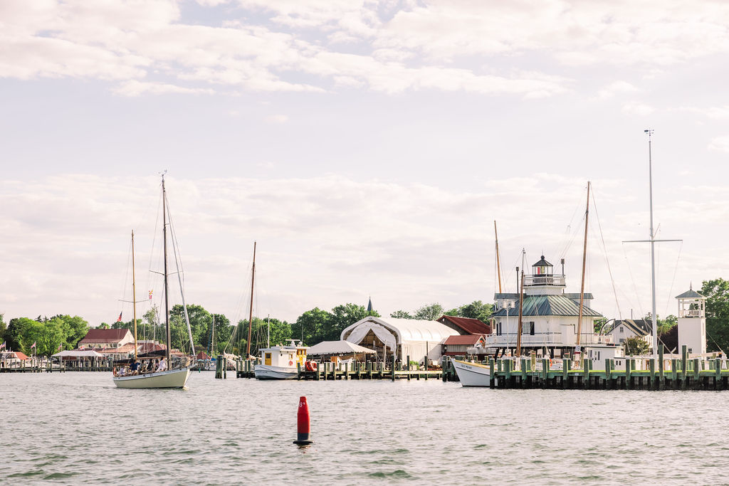 A peaceful harbor scene with sailboats docked at a pier, a white boathouse, and a lighthouse against a cloudy sky, surrounded by green trees and small buildings.