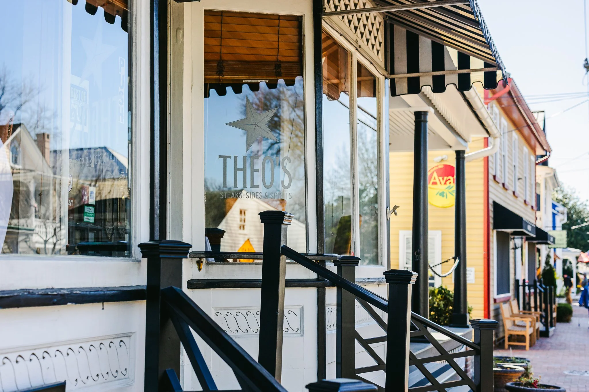 Storefront of THEO'S restaurant with large glass window displaying a star logo and the name, featuring a black and white striped awning, with exterior stairs and sidewalk seating in a small town.