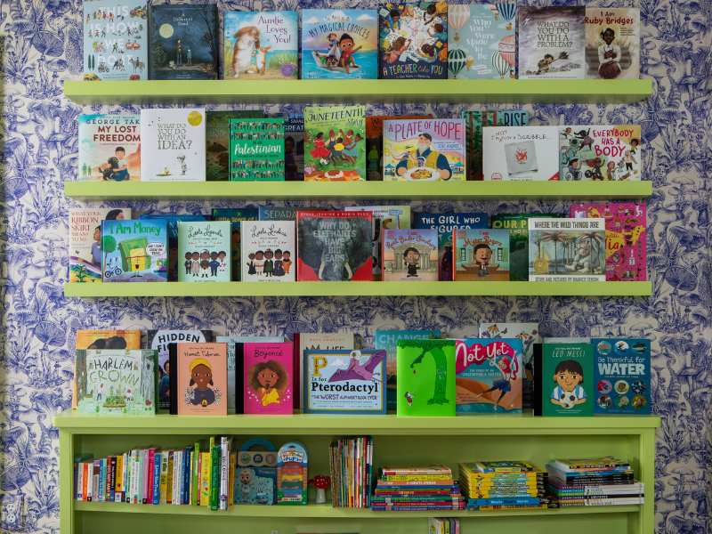 A wall of children's books arranged on green shelves against a blue and white floral wallpaper background. The books include titles about animals, adventures, and educational topics, with some standing upright and others stacked. A lower bookshelf ho