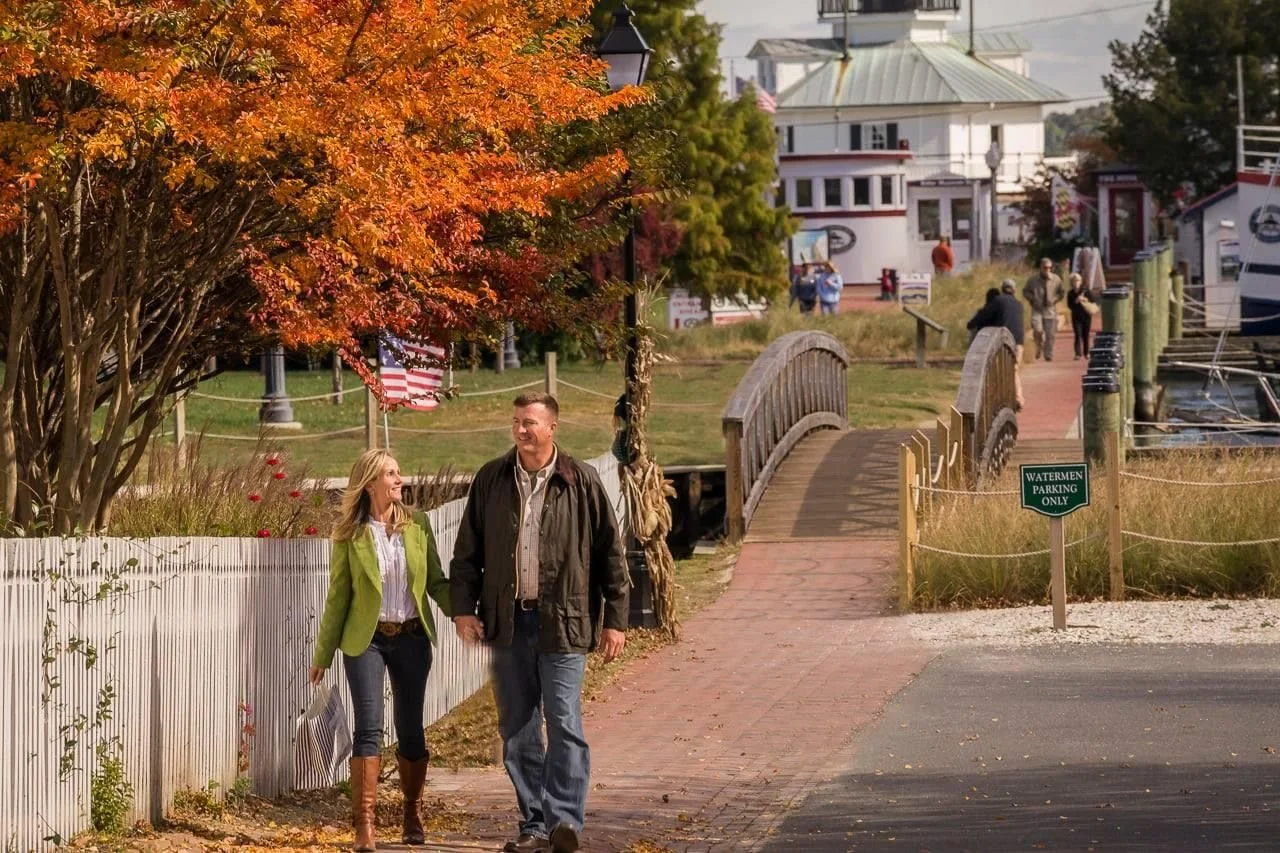 A man and woman walking along a brick pathway by a white picket fence and autumn trees near a waterfront with boats and buildings in the background.