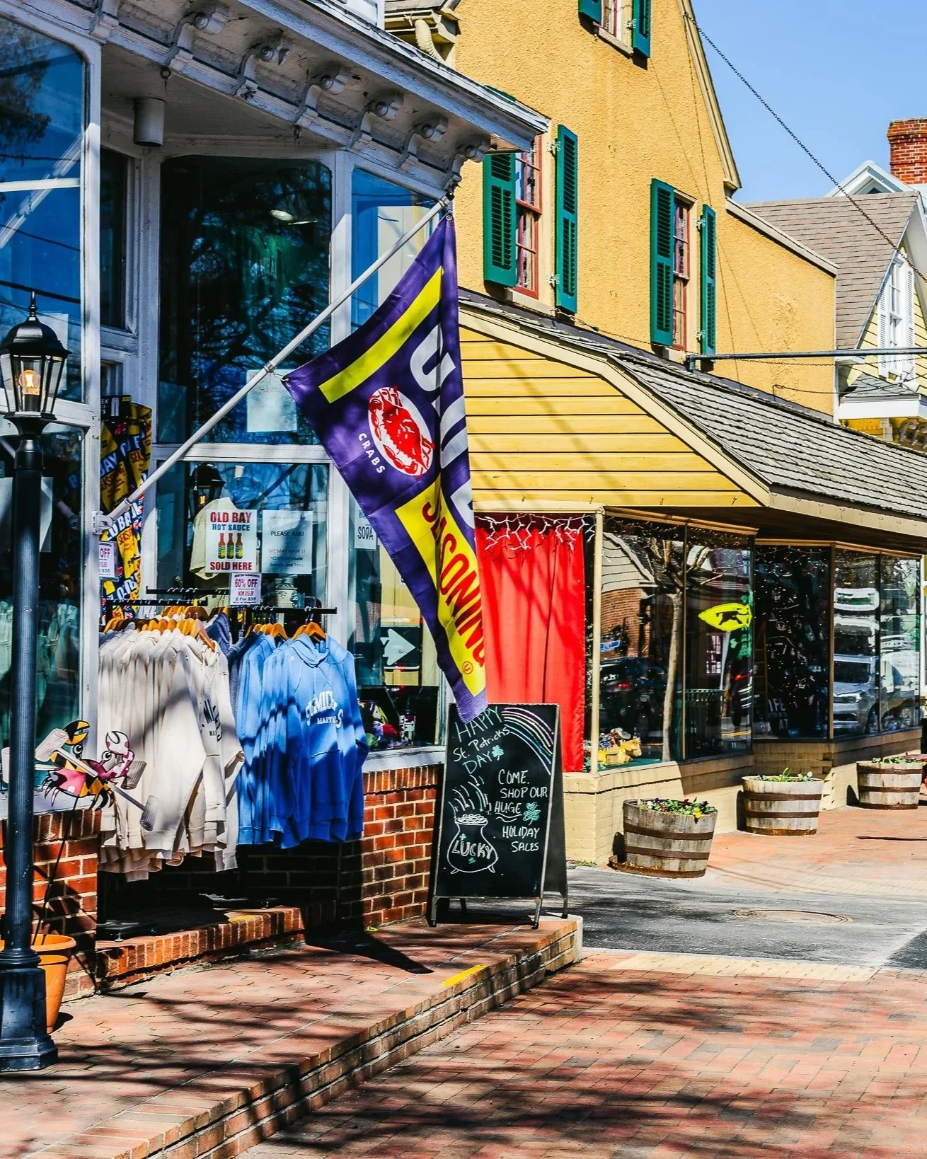 Street view of a small retail shop with a display of clothing on the sidewalk, flags, and a blackboard sign advertising holiday sales and St. Patrick's Day promotions, with yellow and brown buildings and brick pavement.