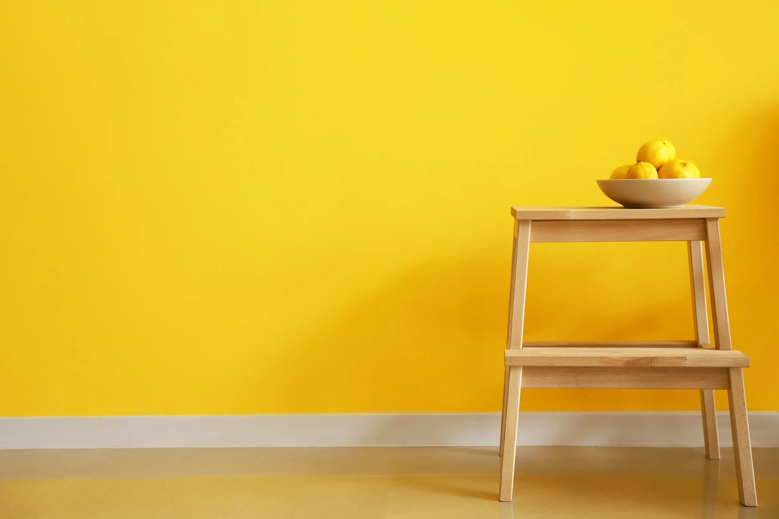 A bowl of yellow lemons sits on a brown stool in front of a bright yellow wall.