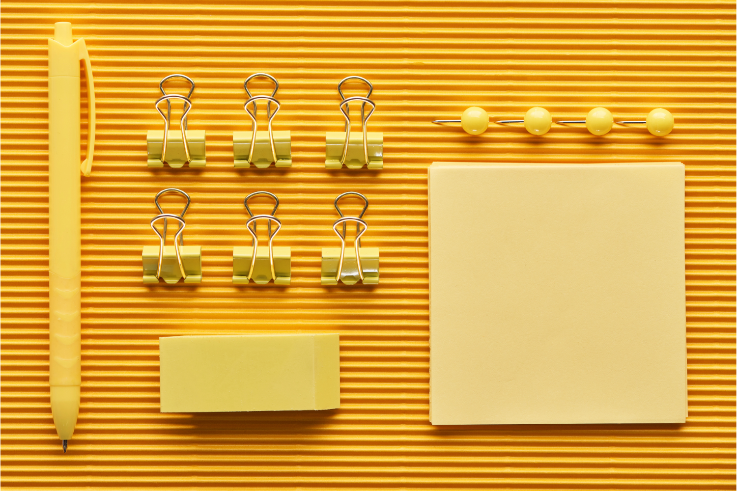 Overhead flat lay of a yellow pin, six clips, two packets of post-its, and four push pins, all yellow, organized on a textured mat.