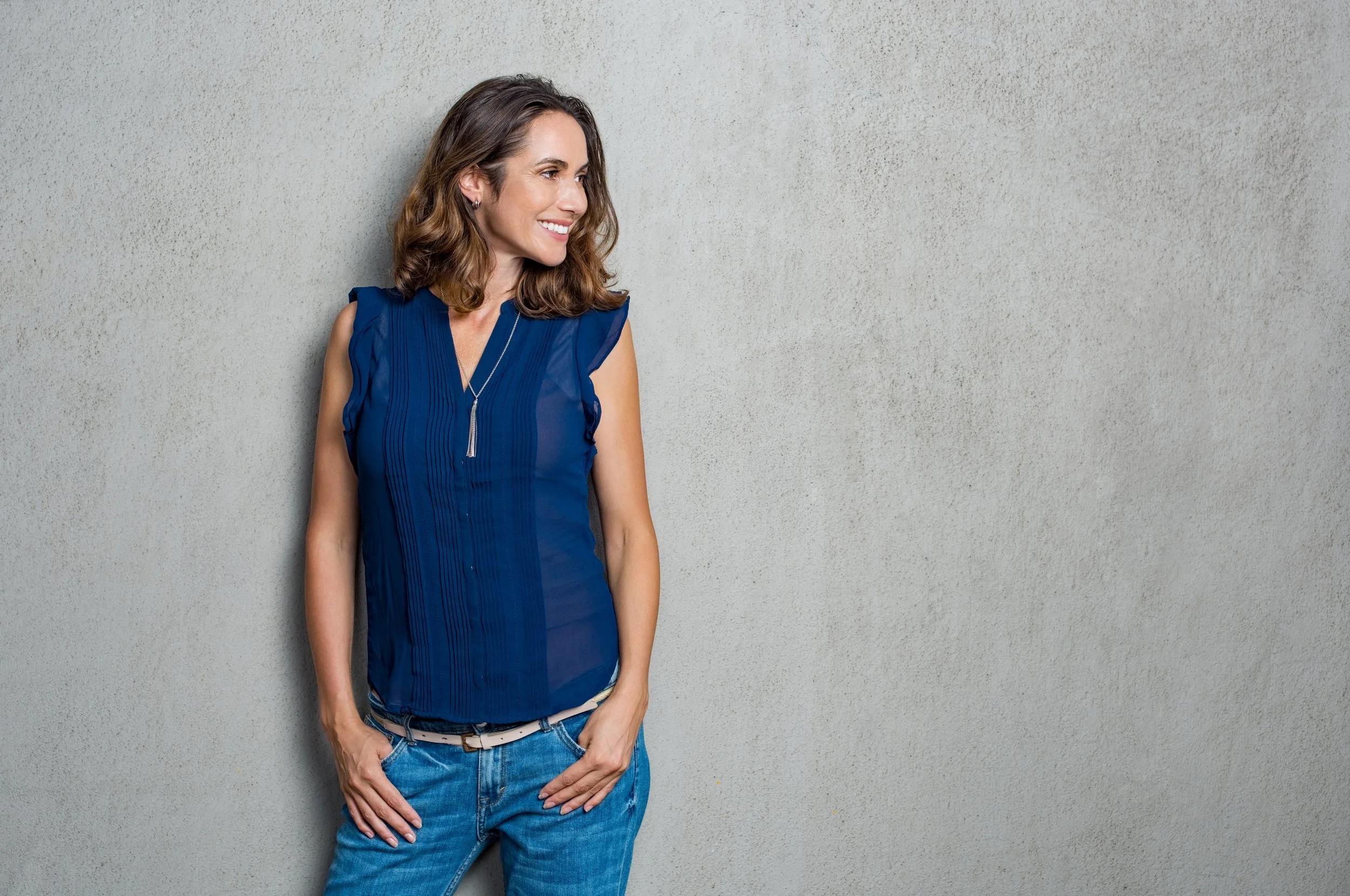 A female small business owner stands in front of a grey wall. Her thumbs are in her jeans pockets and her head is turned, smiling at something off screen.