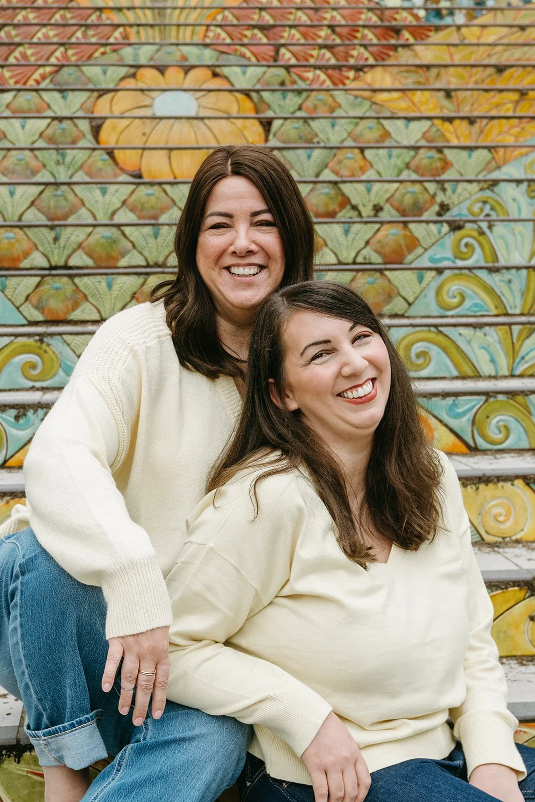 A mother and daughter sit on mosiac steps. The daughter sits lower than her mother, and leans her elbow on her mother's lap, and they both smile at the camera. They are a mother-daughter business duo and this is their headshot.