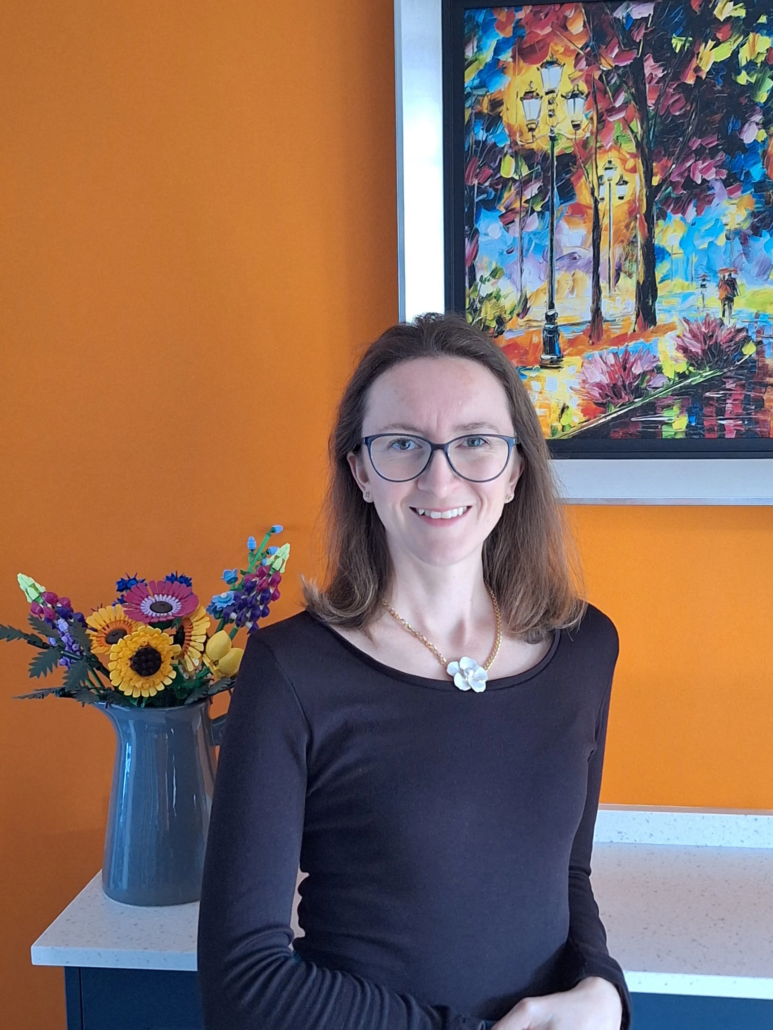 A female small business owner stands against an orange wall, her elbow resting on a counter. A vase of flowers is on the counter behind her as well. She smiles confidently at the camera. 