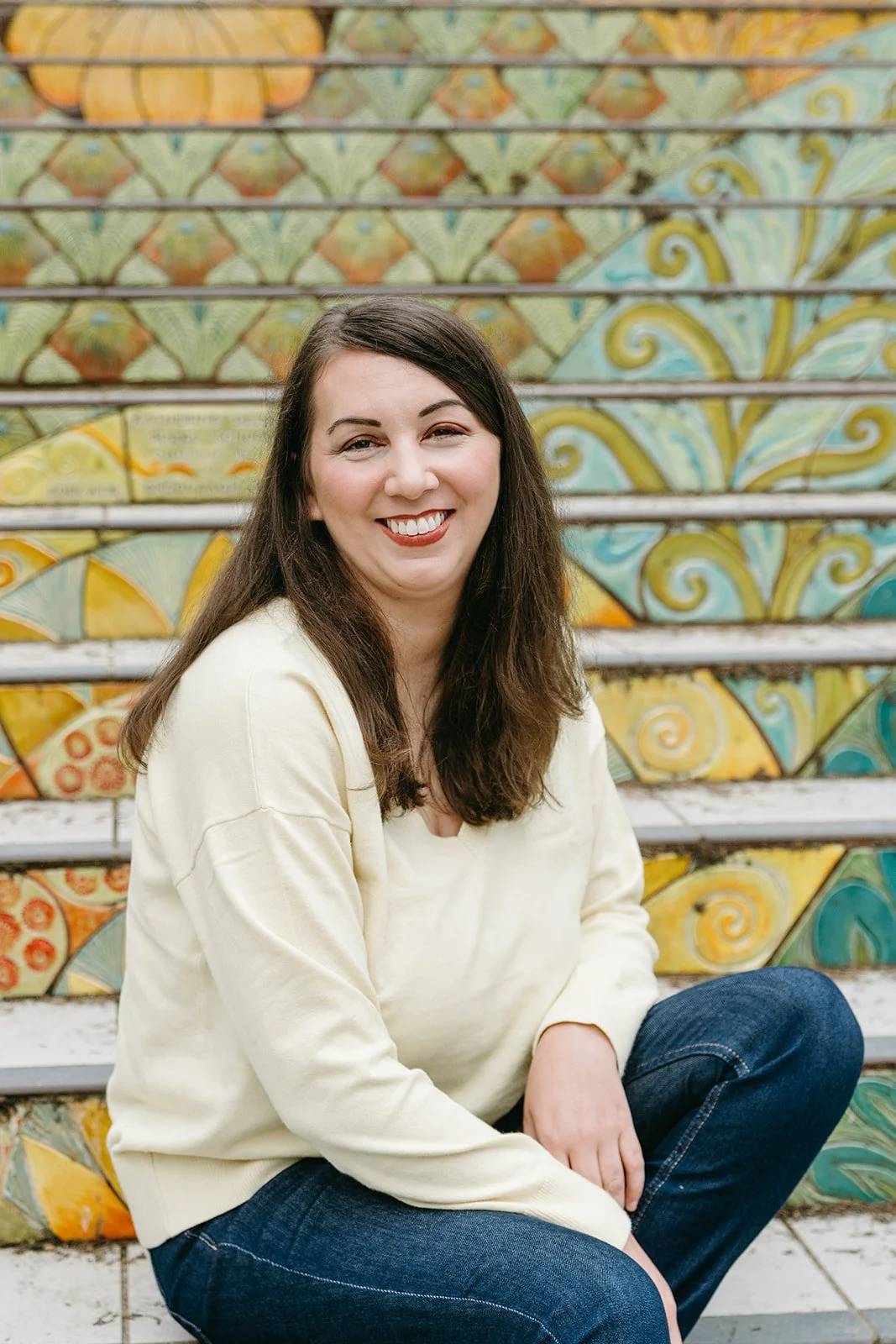 Kate, a Marketing Specialist, sits outdoors on colorful mosiac steps. Her arms are folded in her lap and she turns her head to the camera and smiles.