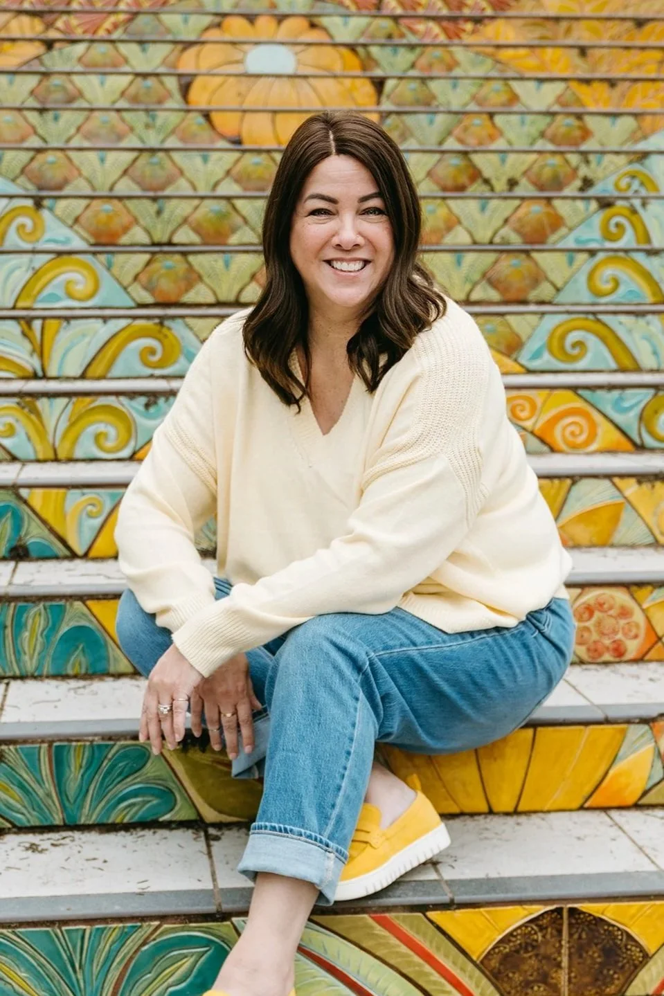 Trish, a Systems Specialist, sits on colorful mosiac steps. She leans her elbow on one knee, toward the camera, and smiles.