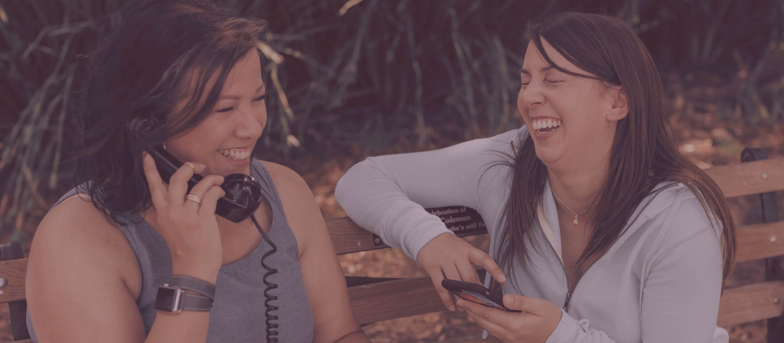 Two women business owners sitting on a park bench, smiling and laughing while one talks on the phone and the other has her cell phone in her hands. They are a mother daughter business duo.