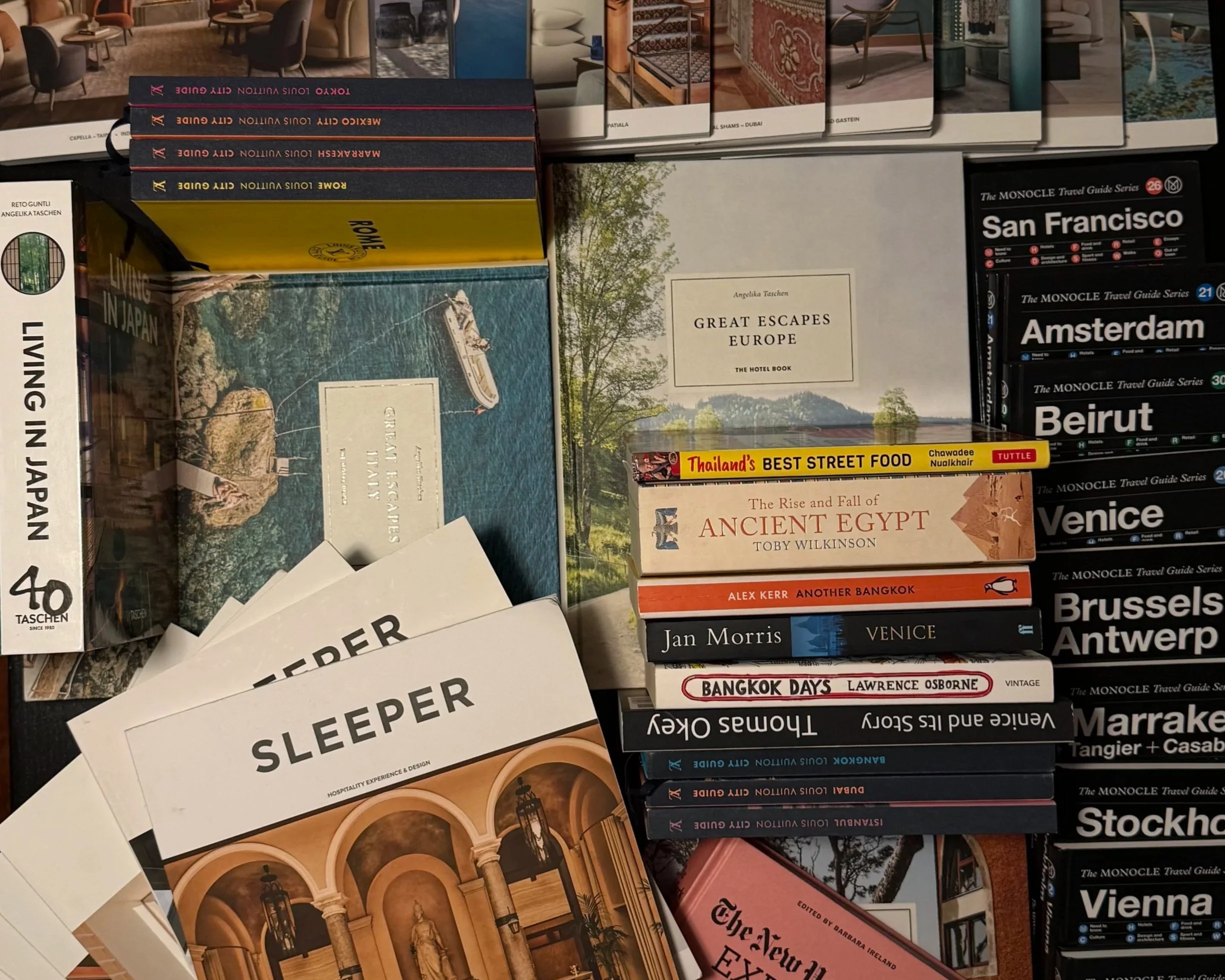 Stack of travel guides, magazines, and brochures featuring destinations like San Francisco, Amsterdam, Beirut, Venice, and Bangkok, along with books on Europe, ancient Egypt, and Japan. Pages display images of interior designs, landmarks, and cityscapes.