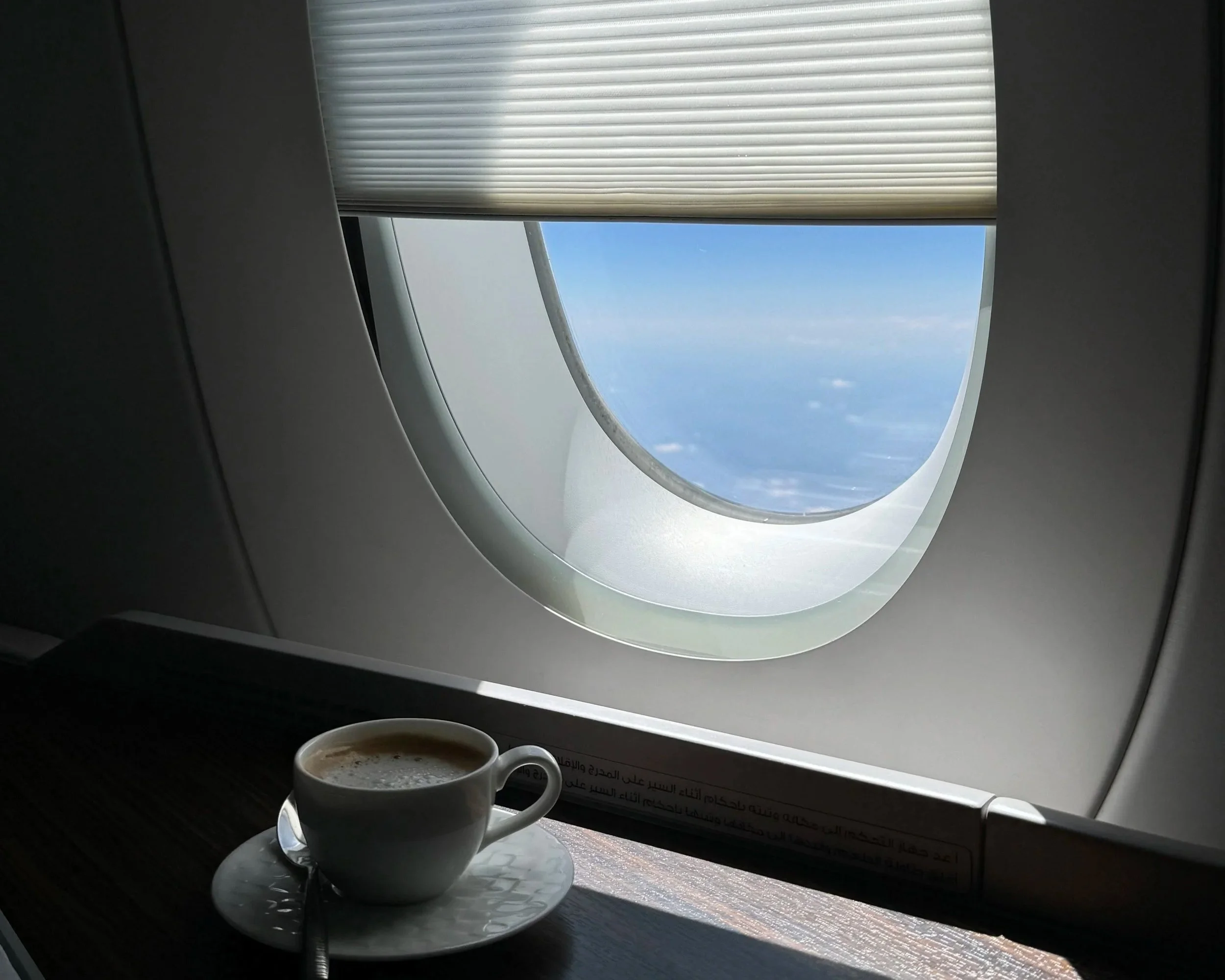 A cup of coffee on a small tray placed on an airplane window ledge. The window shows a view of the blue sky and some clouds, with blinds partially pulled down.