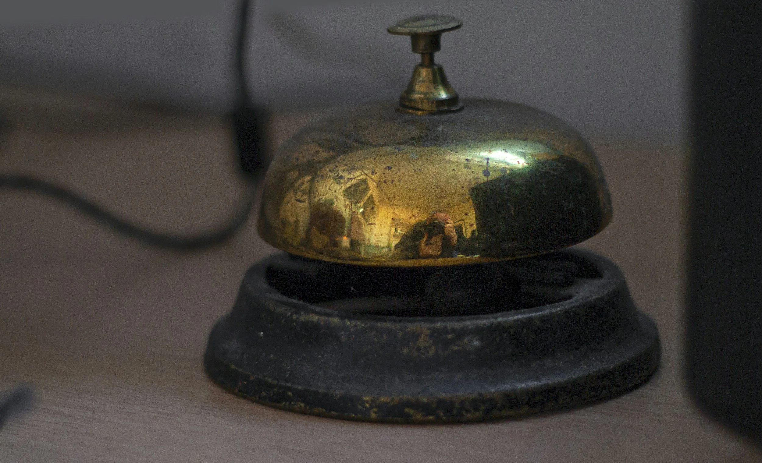 A vintage brass bell with a black base on a wooden surface, reflecting a room interior and a person taking the photo.
